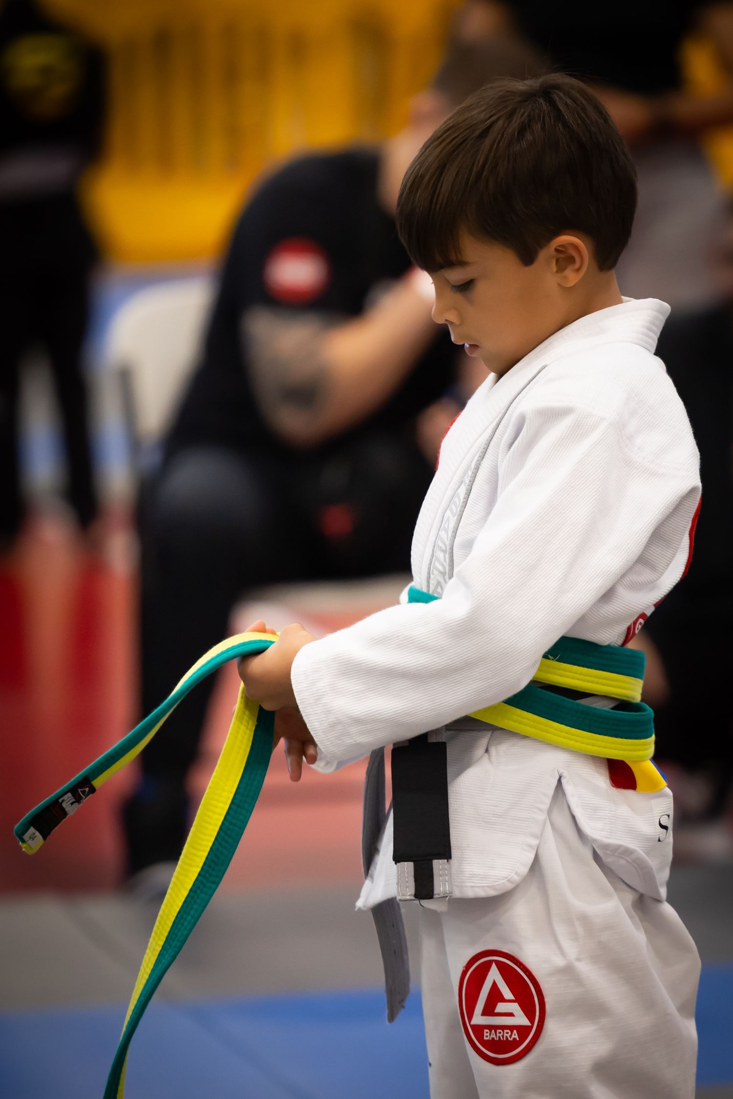 Young boy in white Gracie Barra BJJ gi tying his yellow-green belt at a martial arts tournament.
