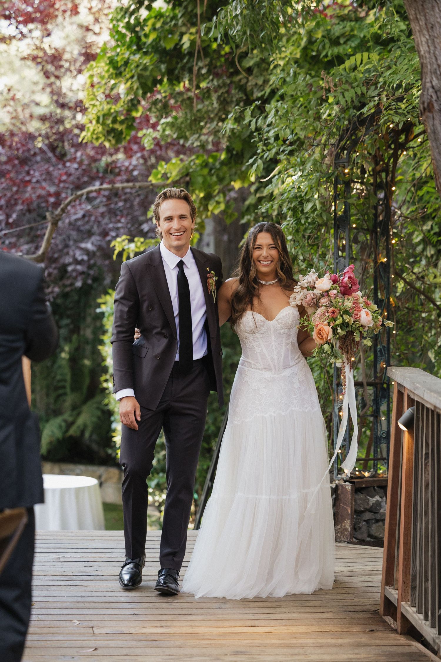Smiling newlywed couple walks down outdoor wooden aisle, bride in white gown holding bouquet, groom in dark suit.
