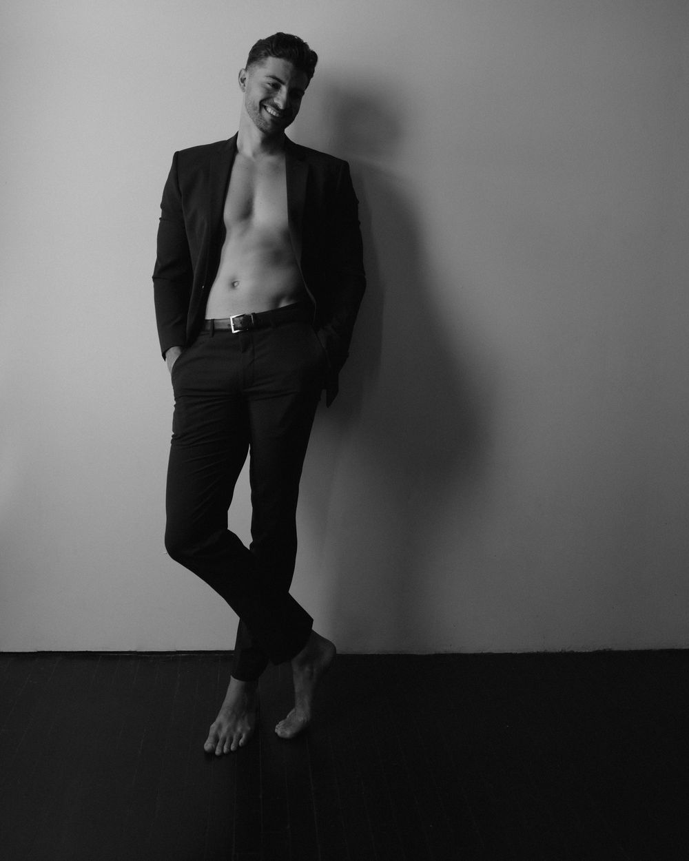 Black and white photo of a shirtless man in suit jacket and trousers, smiling confidently against a wall.