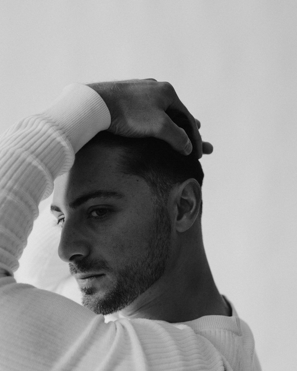 Black and white portrait of a young man in a white sweater, hand resting on his head, looking away.