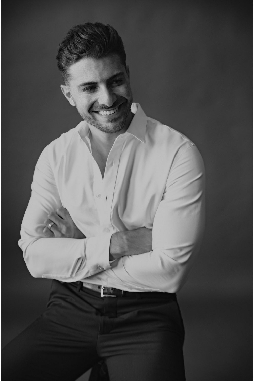 Smiling man with crossed arms wearing white dress shirt and dark pants in black and white studio portrait.