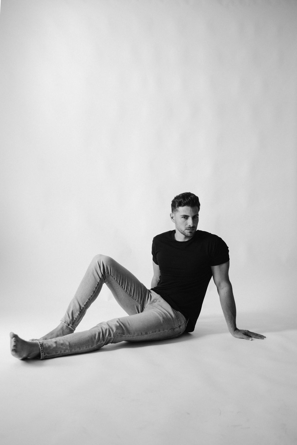 Black and white photo of a young man in a black t-shirt and light jeans sitting on a white studio floor.
