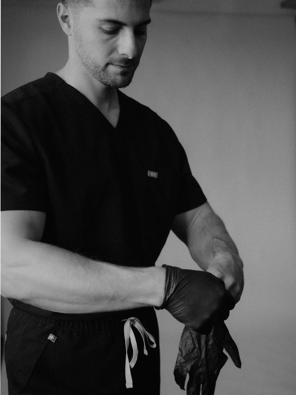 A male healthcare professional in black scrubs puts on dark medical gloves in a black and white photo.