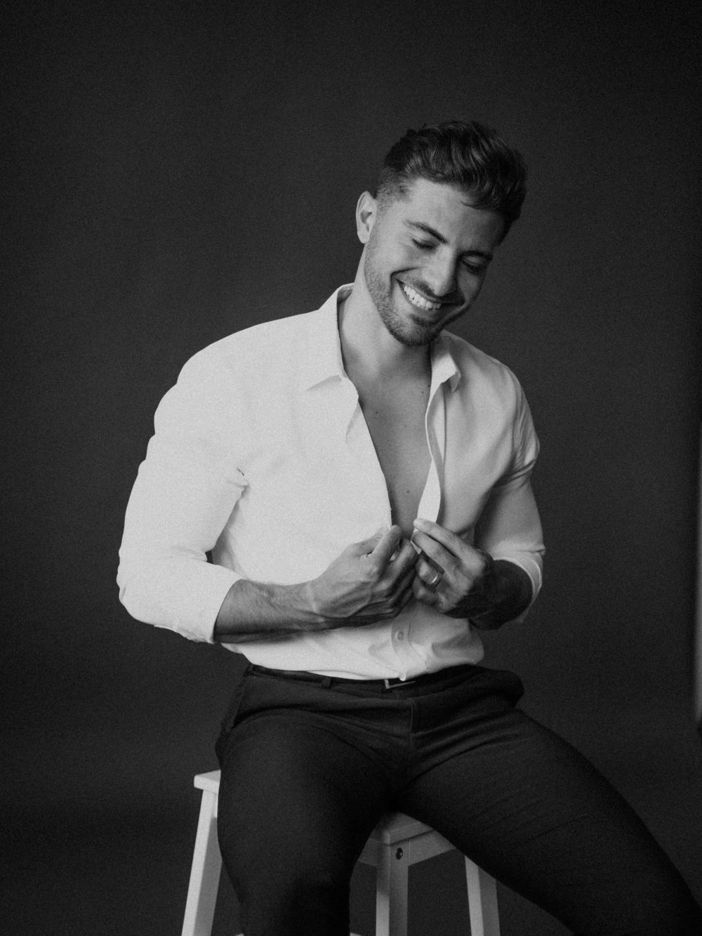 Black and white studio photo of smiling man in white shirt sitting on stool, buttoning shirt.