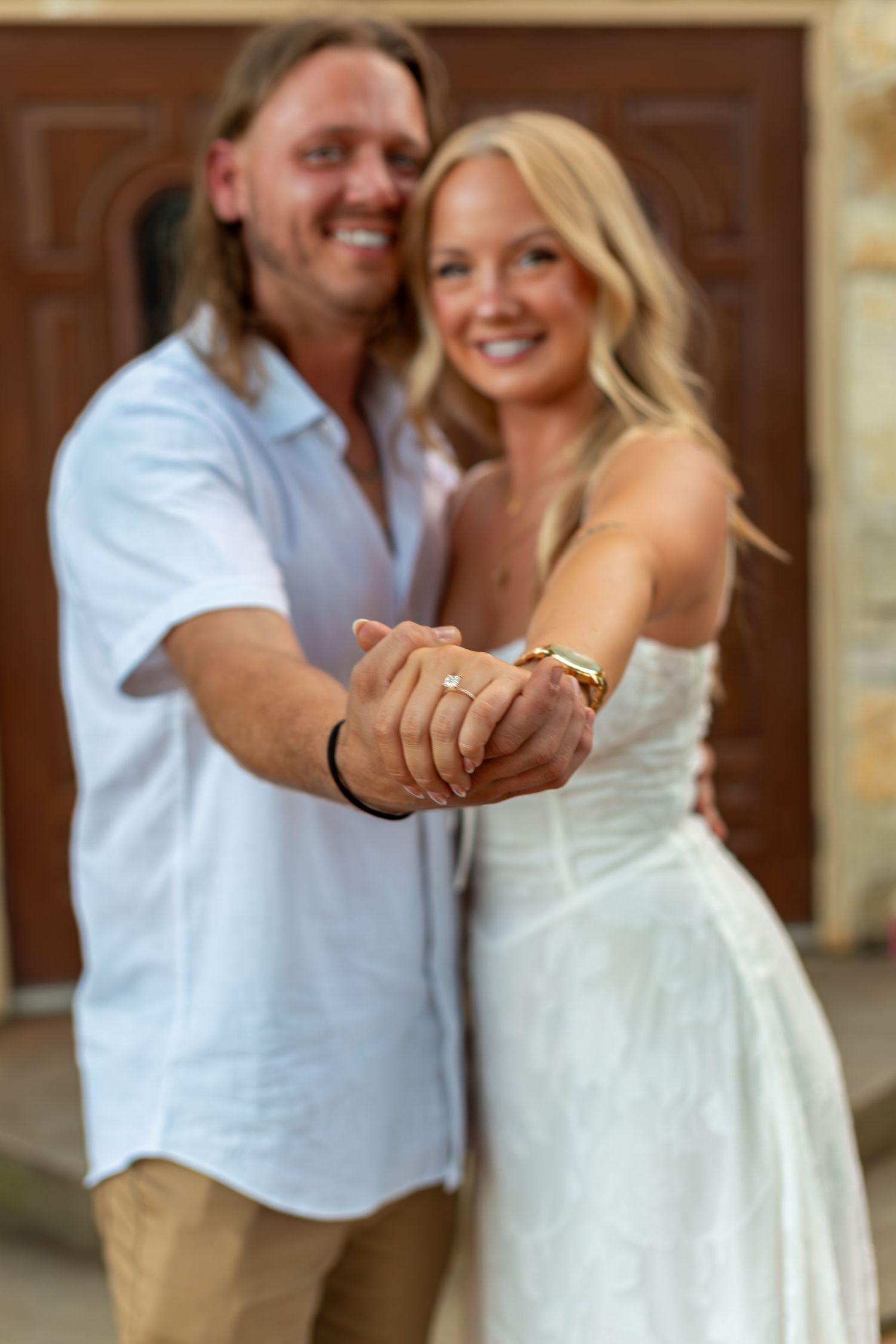 Happy couple showing off engagement ring, man in white shirt embracing blonde woman in white dress outdoors.