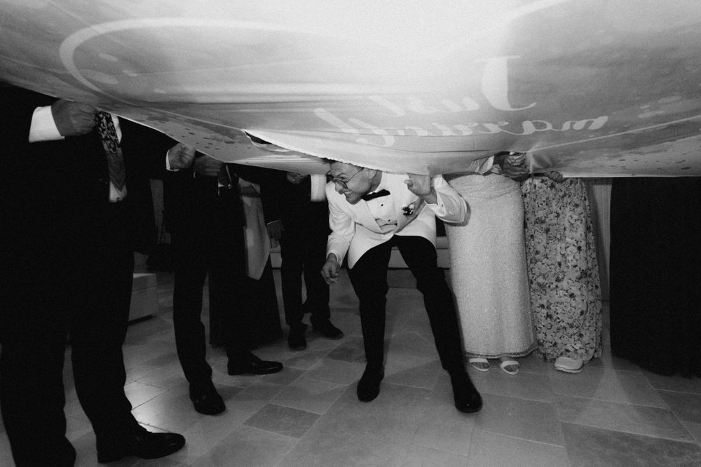 Guests duck under a large white fabric at a lively wedding reception in black and white photo.