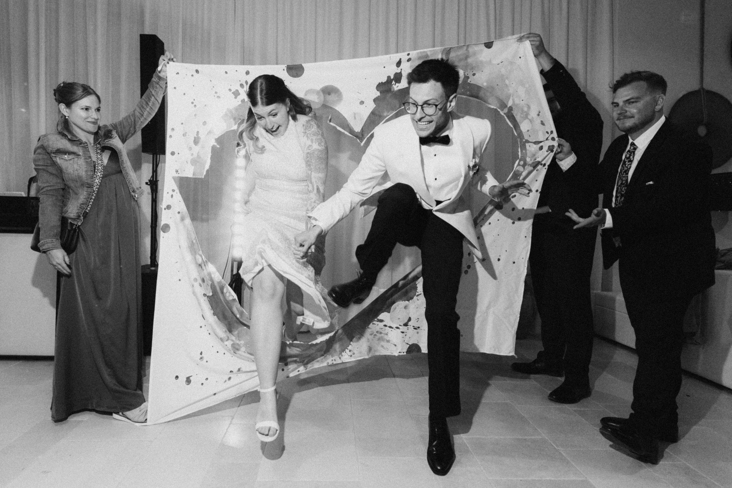 Black and white photo of a bride and groom dancing joyfully in front of a large abstract painting at their wedding reception.