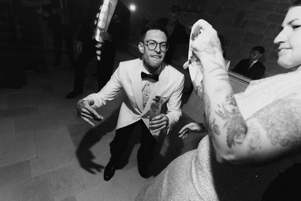 Groom in white tuxedo dancing energetically at wedding reception in black and white photo.