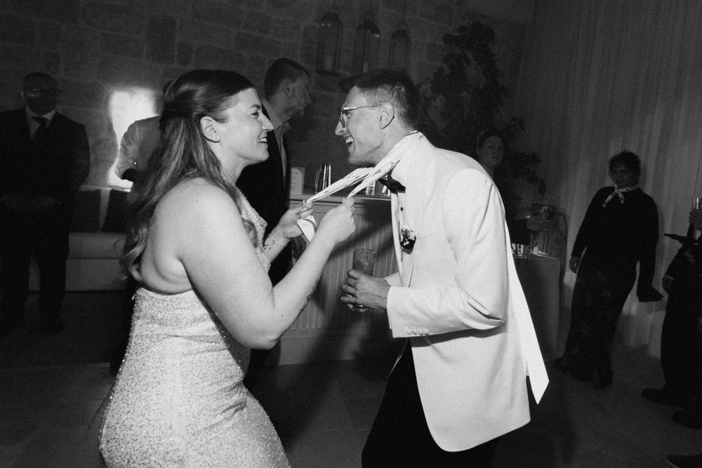 Black and white photo of a couple dancing joyfully at a wedding reception, bride in sequin dress, groom in white tuxedo jacket.