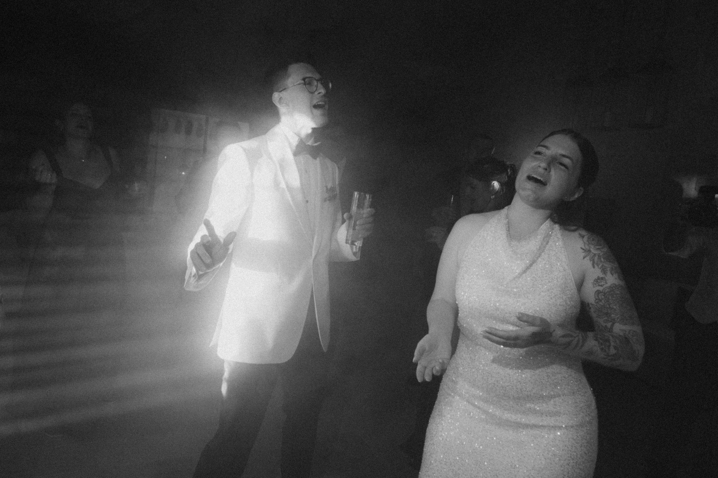 Black and white photo of bride and groom dancing joyfully at their wedding reception under bright lights.