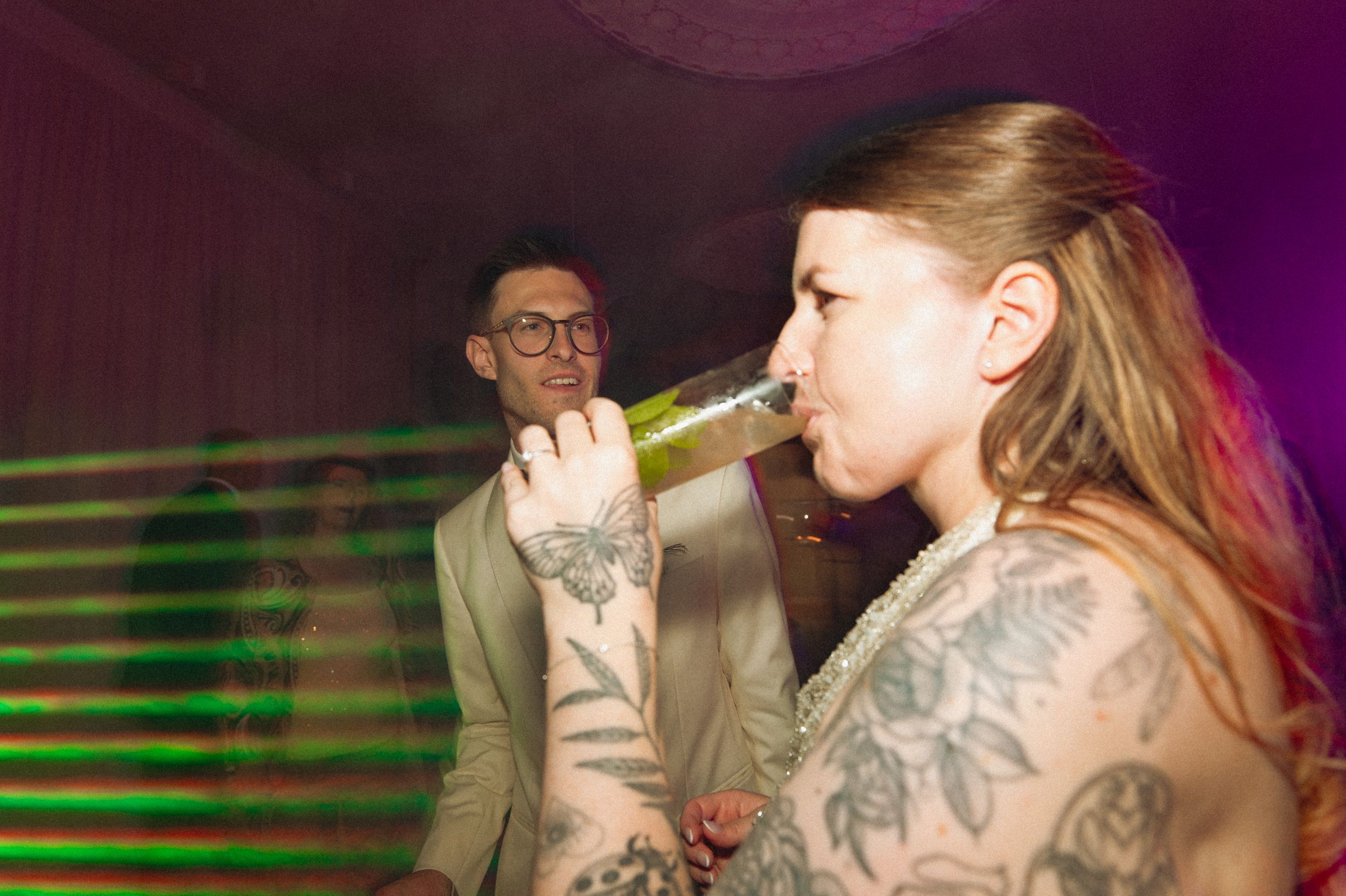 Tattooed woman drinking green beverage at party with man in glasses smiling in background.