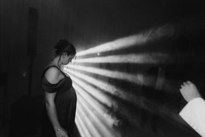 Black and white photo of a woman in a dark room illuminated by dramatic light rays streaming from the side.