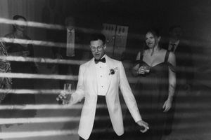 Black and white wedding photo of groom in white tuxedo holding champagne glass with guests celebrating in background.