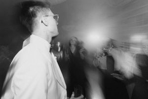 Black and white photo of a man in glasses at a party with blurred guests and bright flash in background.