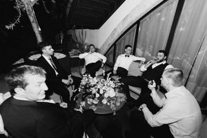 Black and white photo of groomsmen relaxing together around a floral centerpiece at a wedding reception.