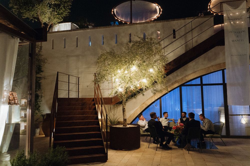 Elegant outdoor patio at night with illuminated tree, staircase, and guests dining under warm string lights.
