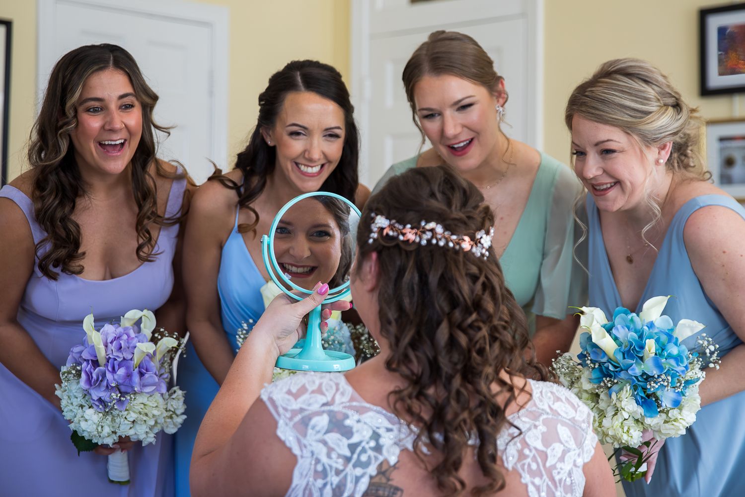 Bride holding mirror surrounded by smiling bridesmaids in blue dresses holding floral bouquets during wedding preparations.
