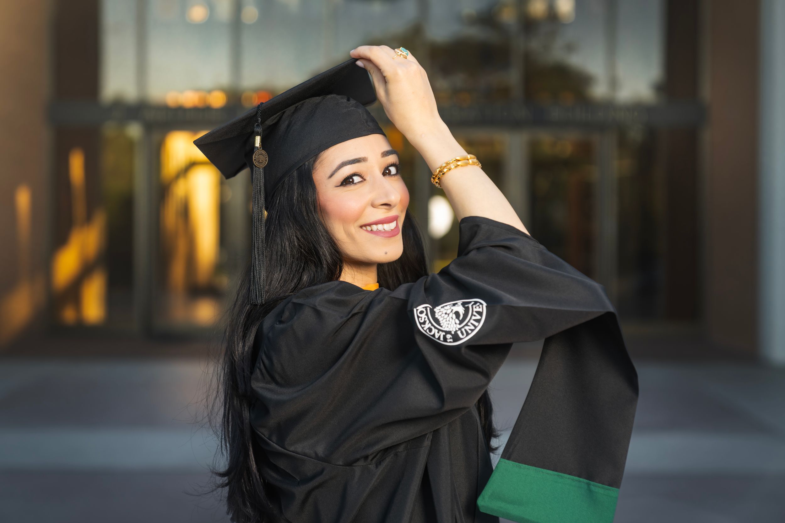 Smiling woman in black graduation cap and gown holding her hat outdoors at a university campus.