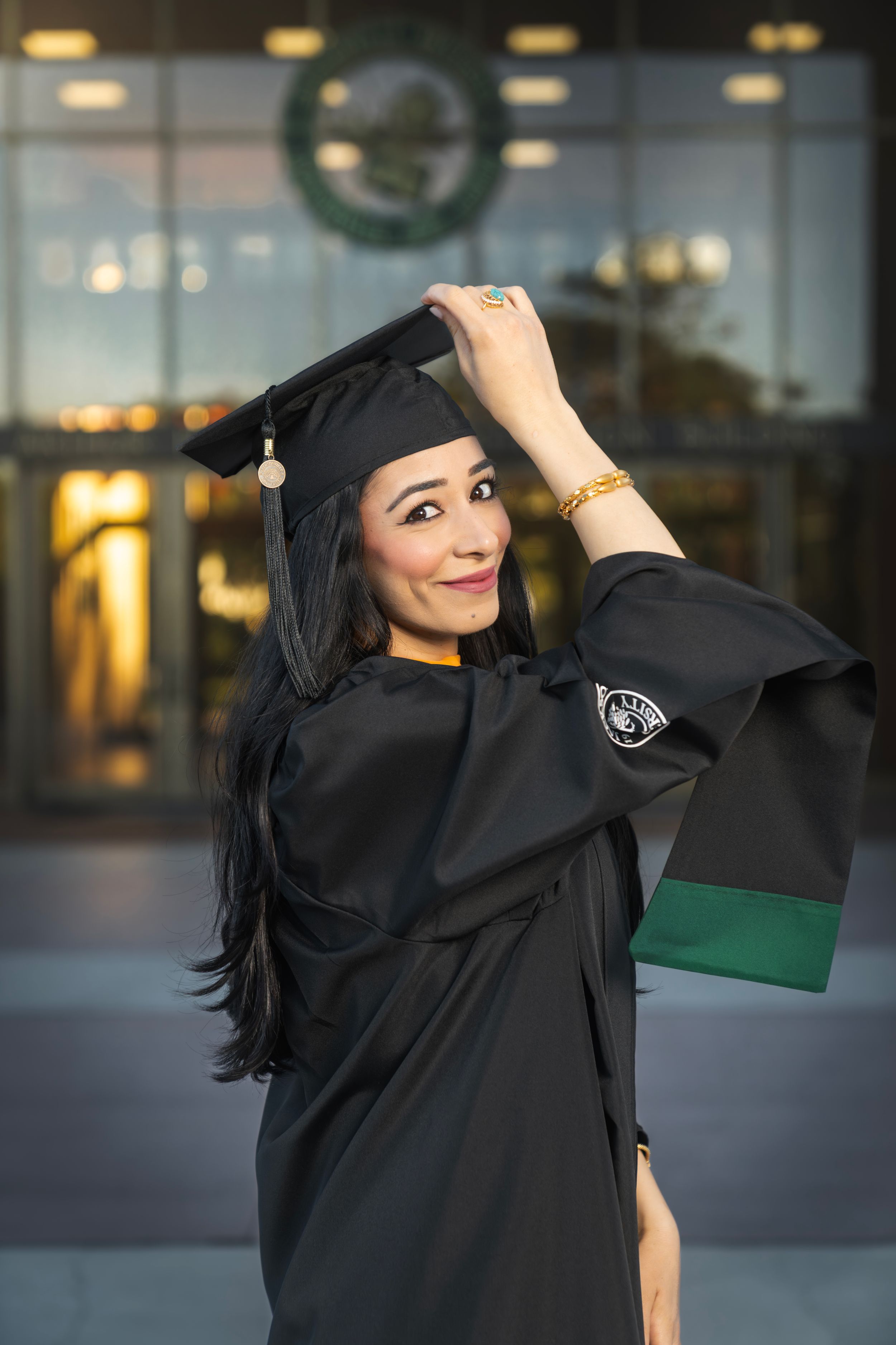 A woman in black graduation cap and gown poses proudly outside a university building with green accents.