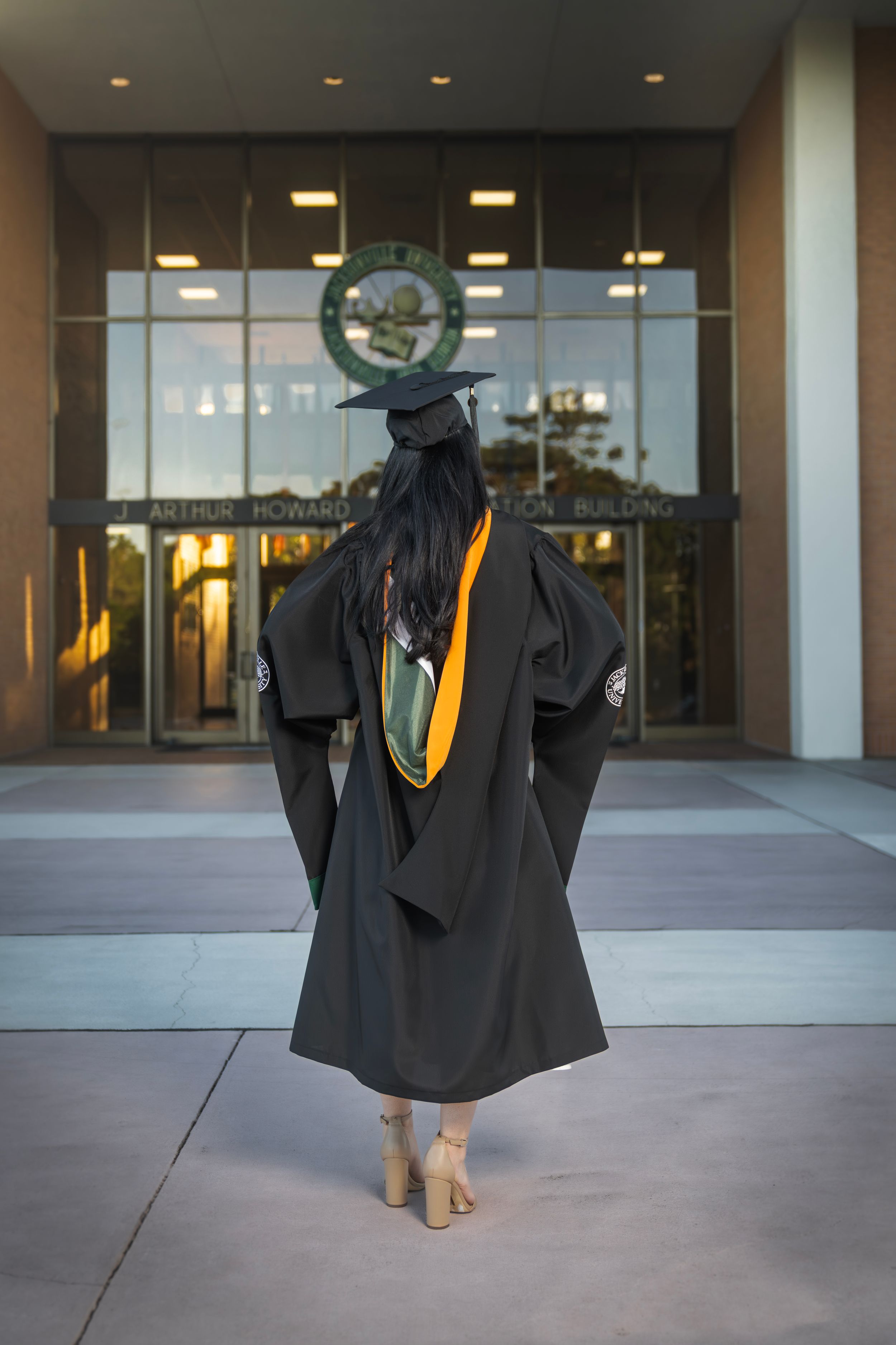 Graduate in black cap and gown stands before Arthur Howard Administration Building entrance.