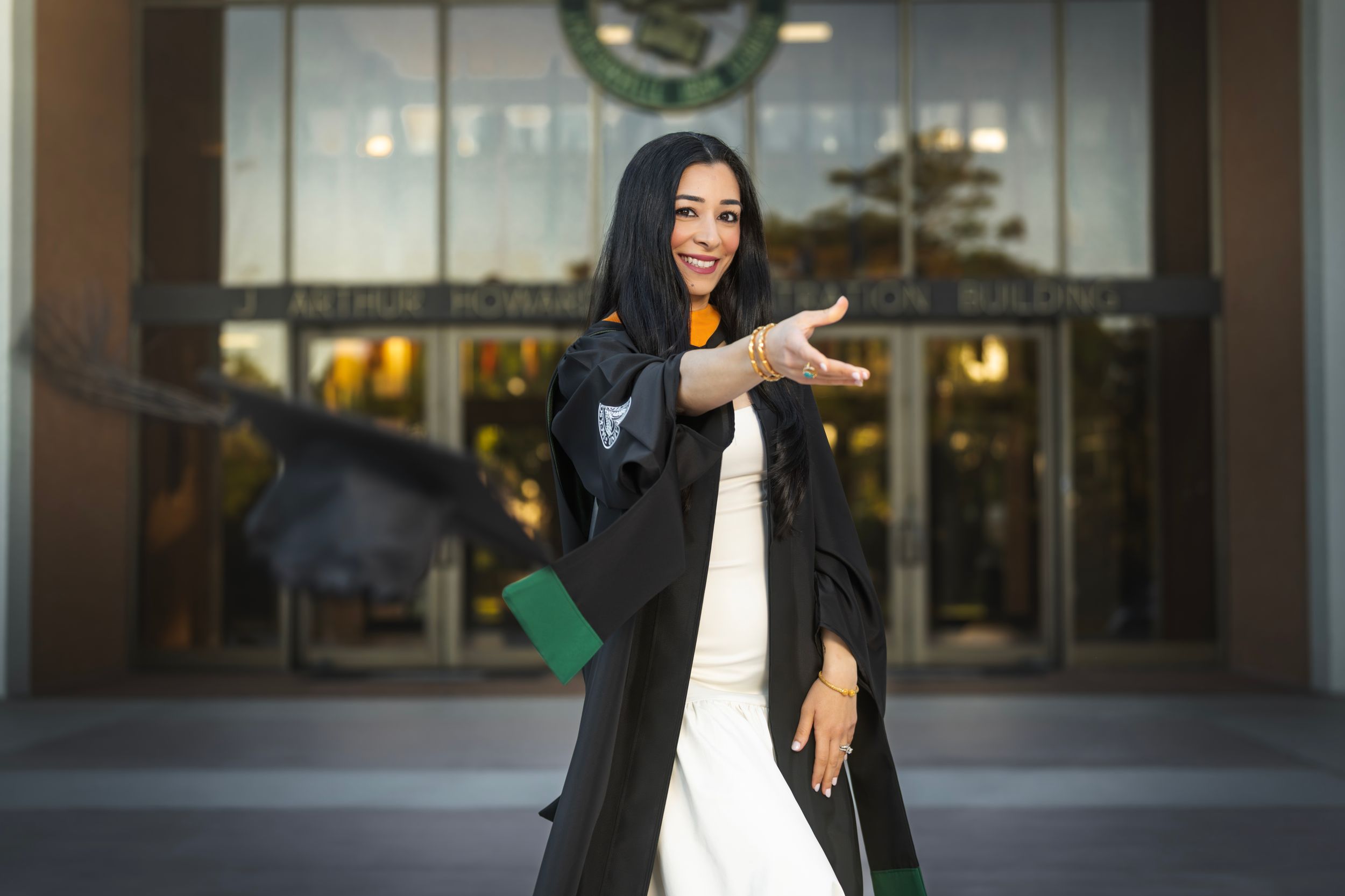 Smiling female graduate in black gown and hijab posing outdoors in front of university building on graduation day.