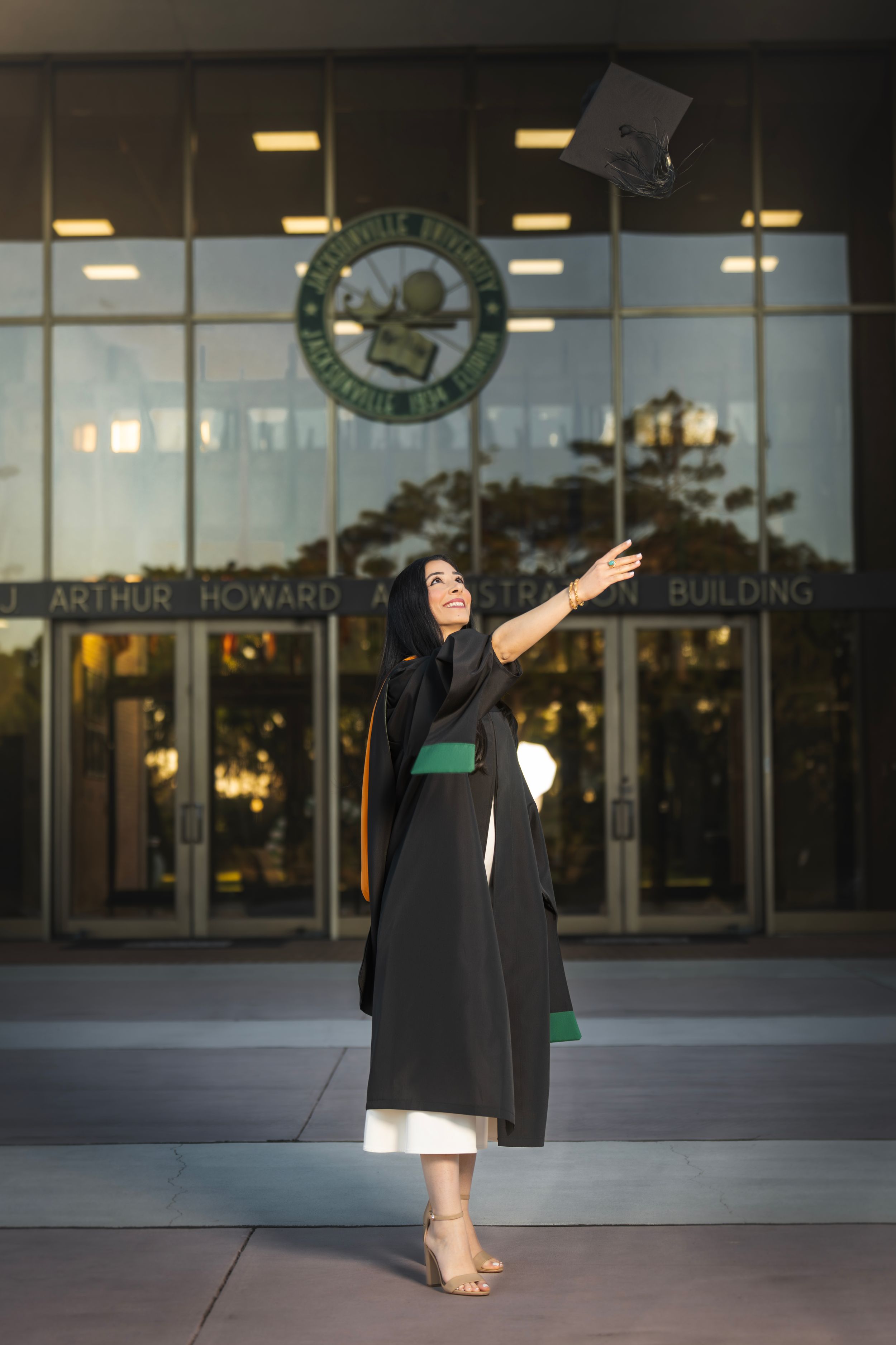 Graduate in black gown tosses cap in front of J. Arthur Howard building, celebrating commencement day.