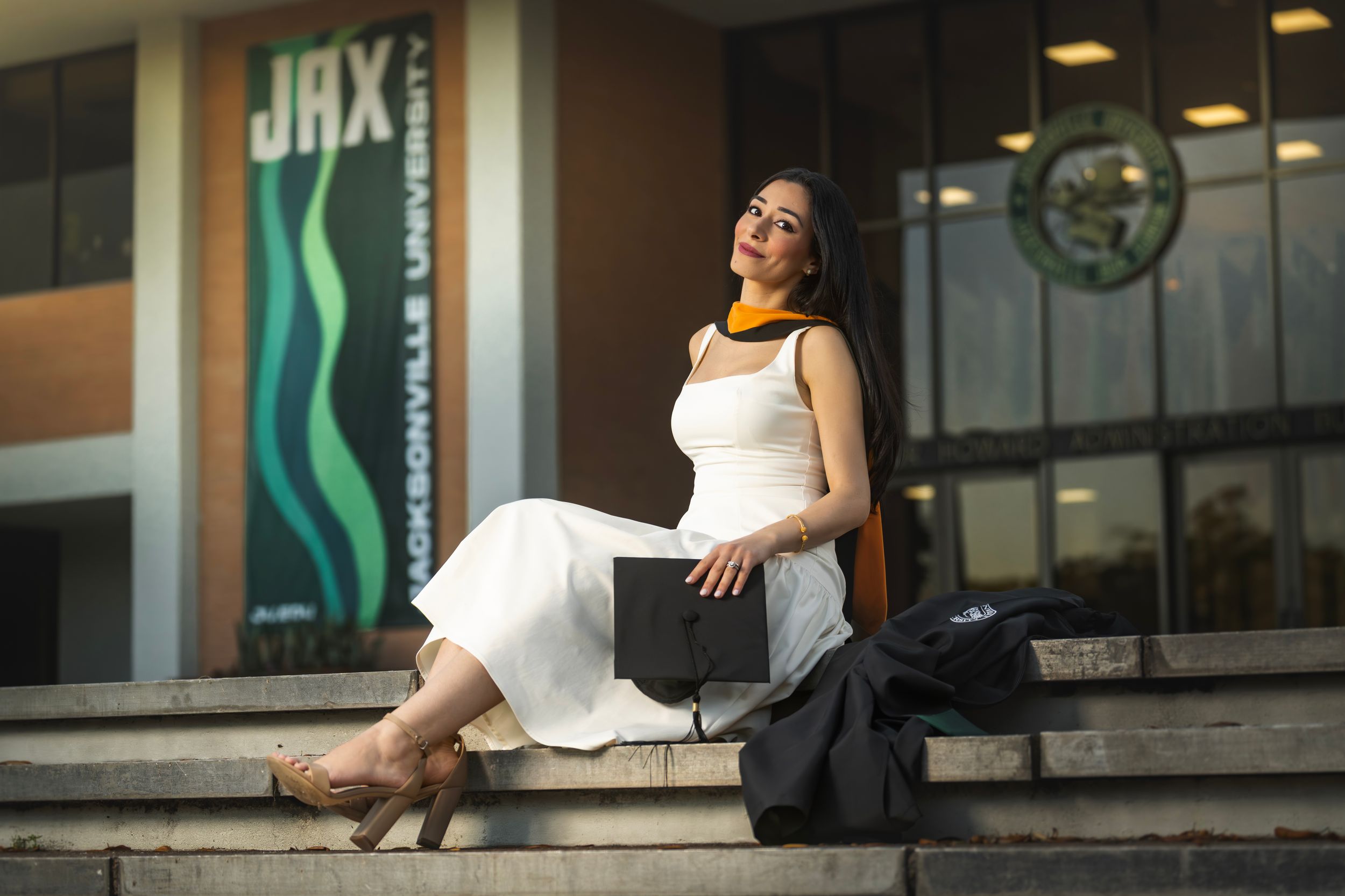Woman in white dress and graduation regalia sits on steps outside Jacksonville University, holding a diploma cover.