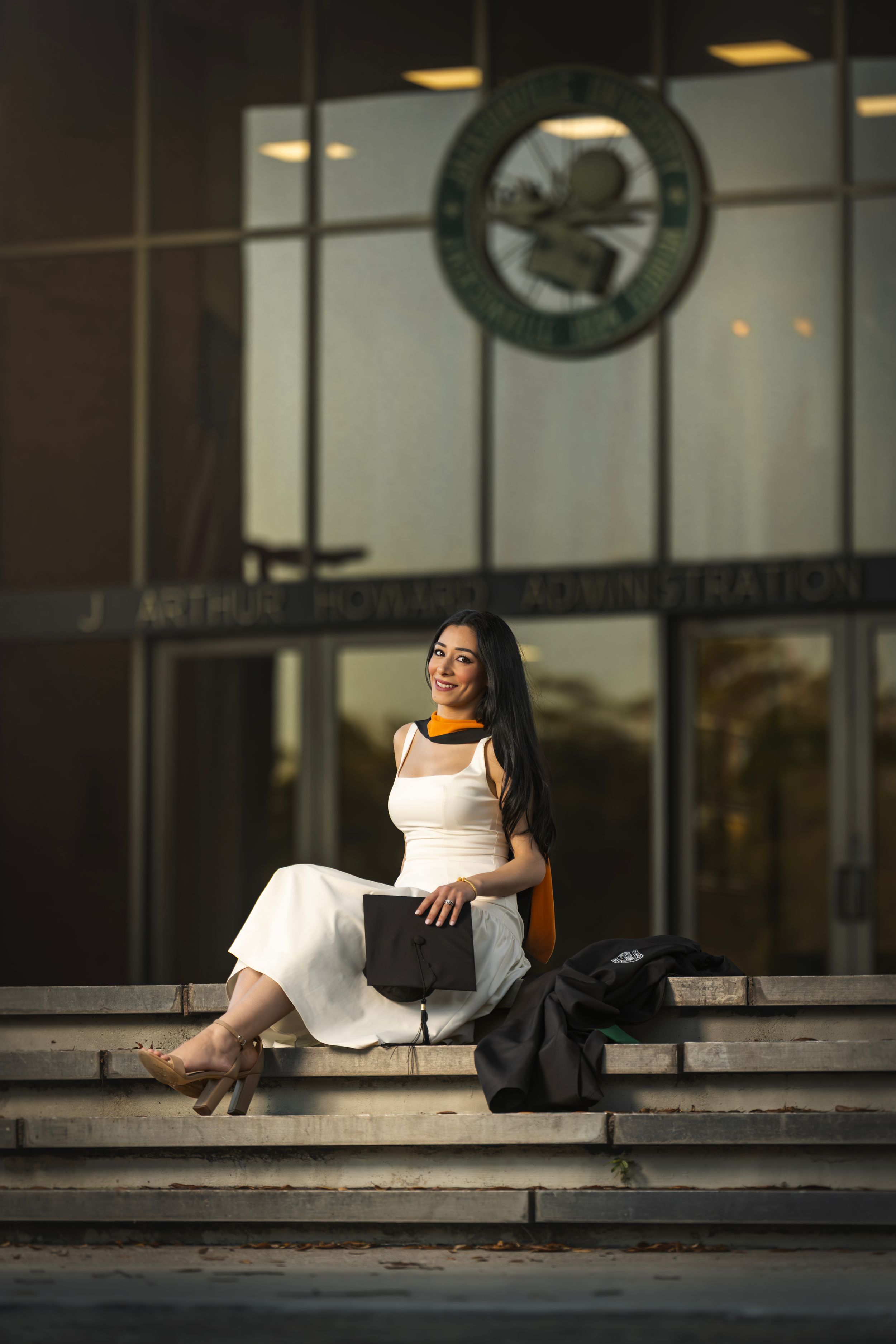 A young woman in a white dress sits on steps outside a university administration building, holding a graduation cap.