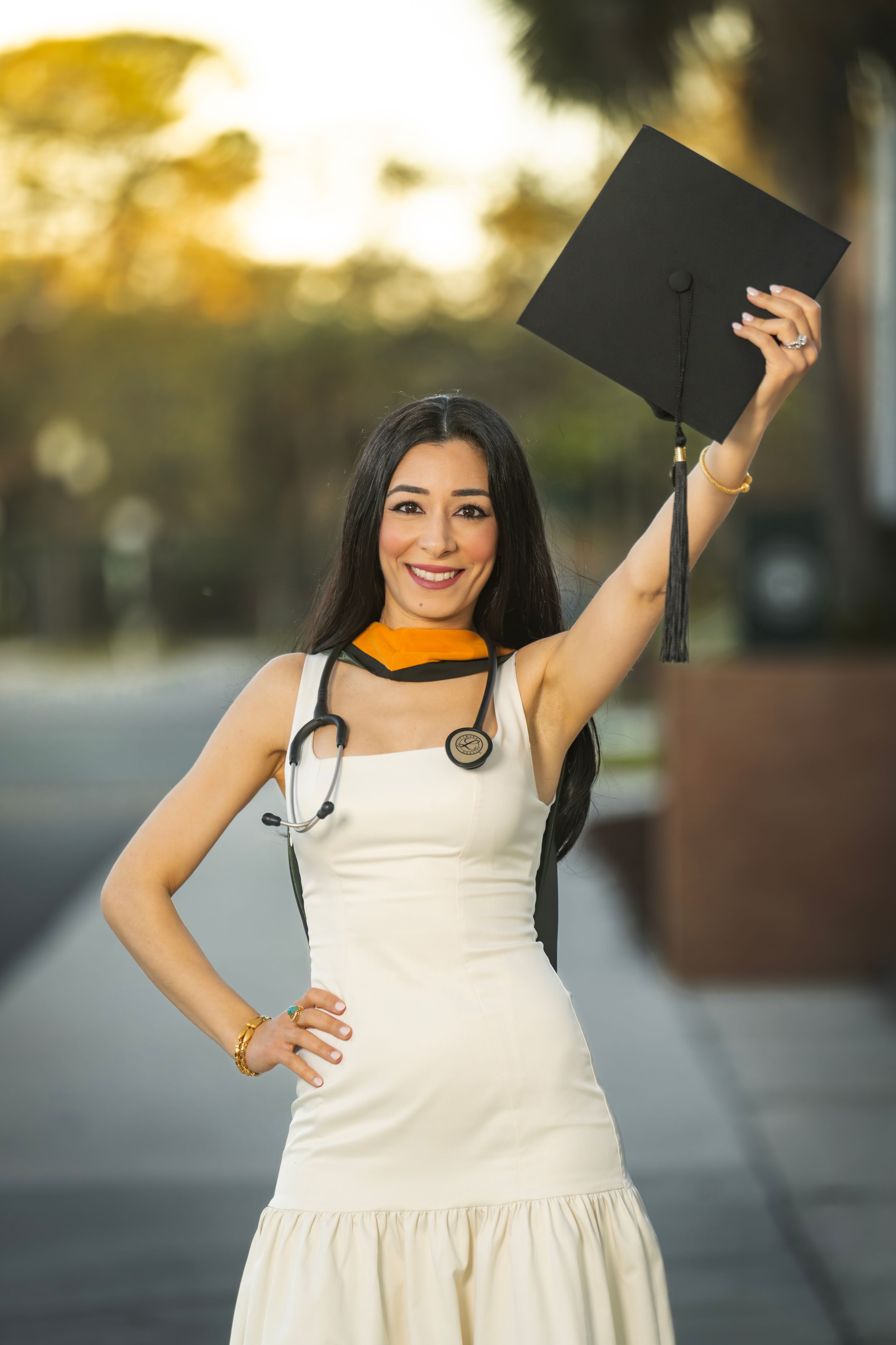 Female medical graduate in white dress holding mortarboard cap with stethoscope, celebrating graduation outdoors.