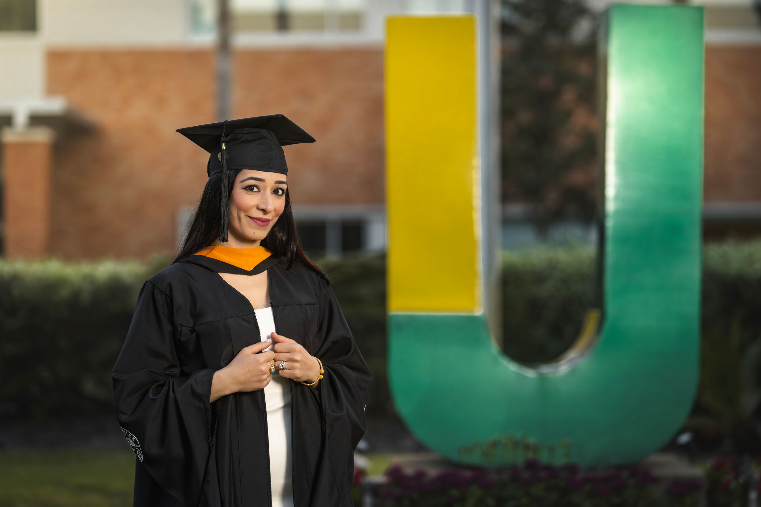 Smiling female graduate in cap and gown poses beside large U university sign outdoors.