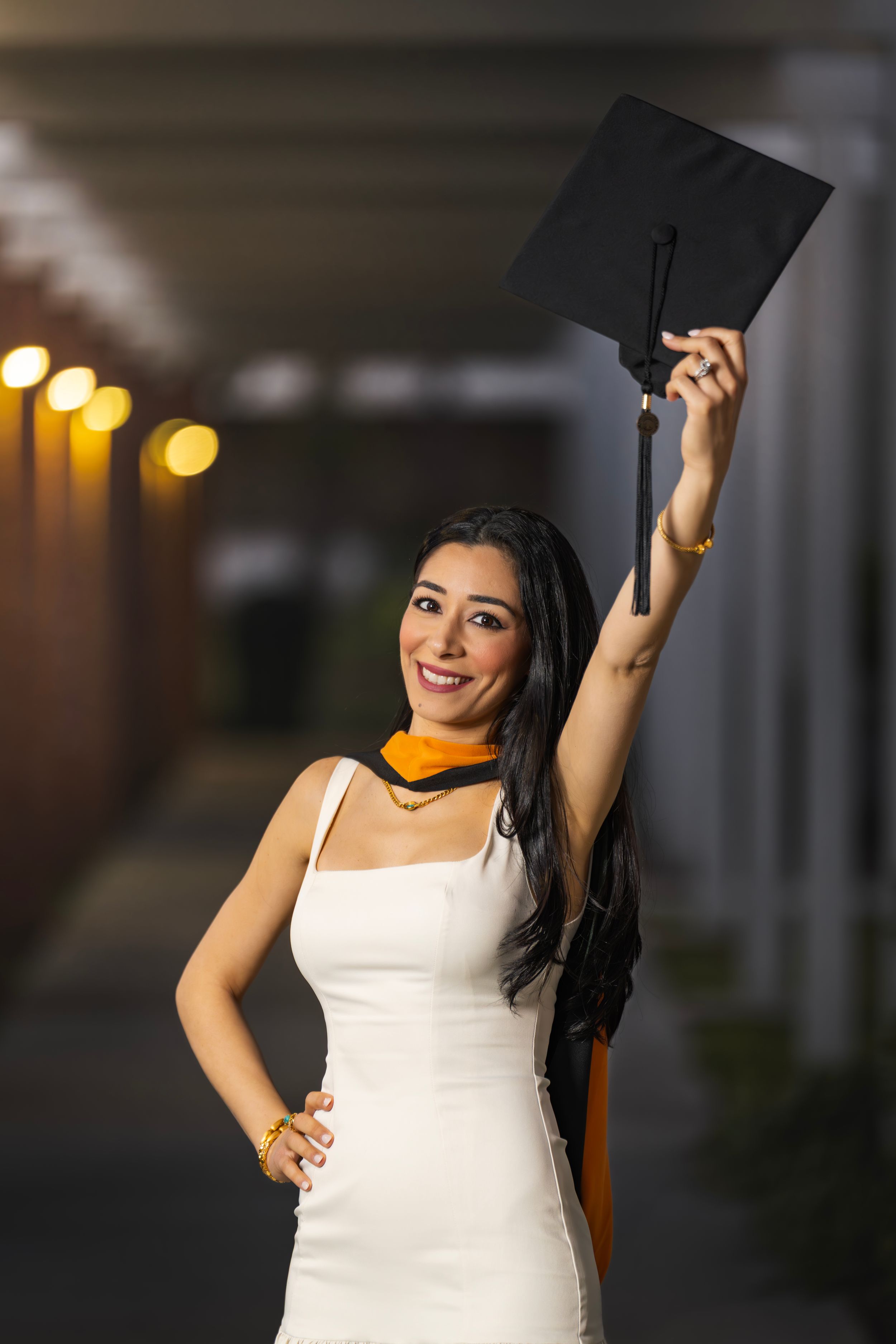 Young woman in white dress holding graduation cap aloft, wearing orange sash, smiling proudly in dimly lit hallway.