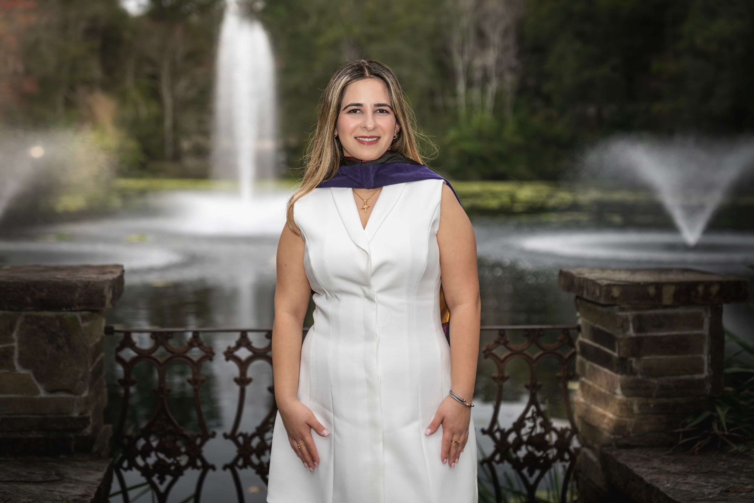 Woman in white dress and purple scarf smiling in front of decorative fountains and iron fence outdoors.