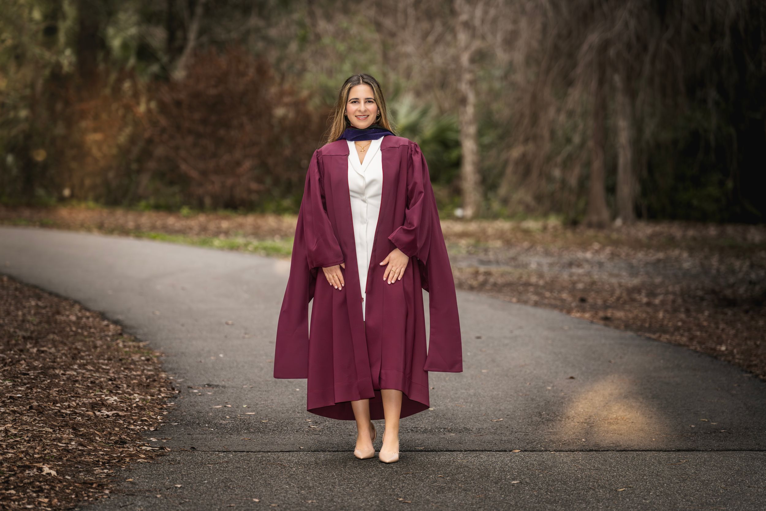 Woman in maroon graduation gown walking on a tree-lined path, smiling confidently on her graduation day.