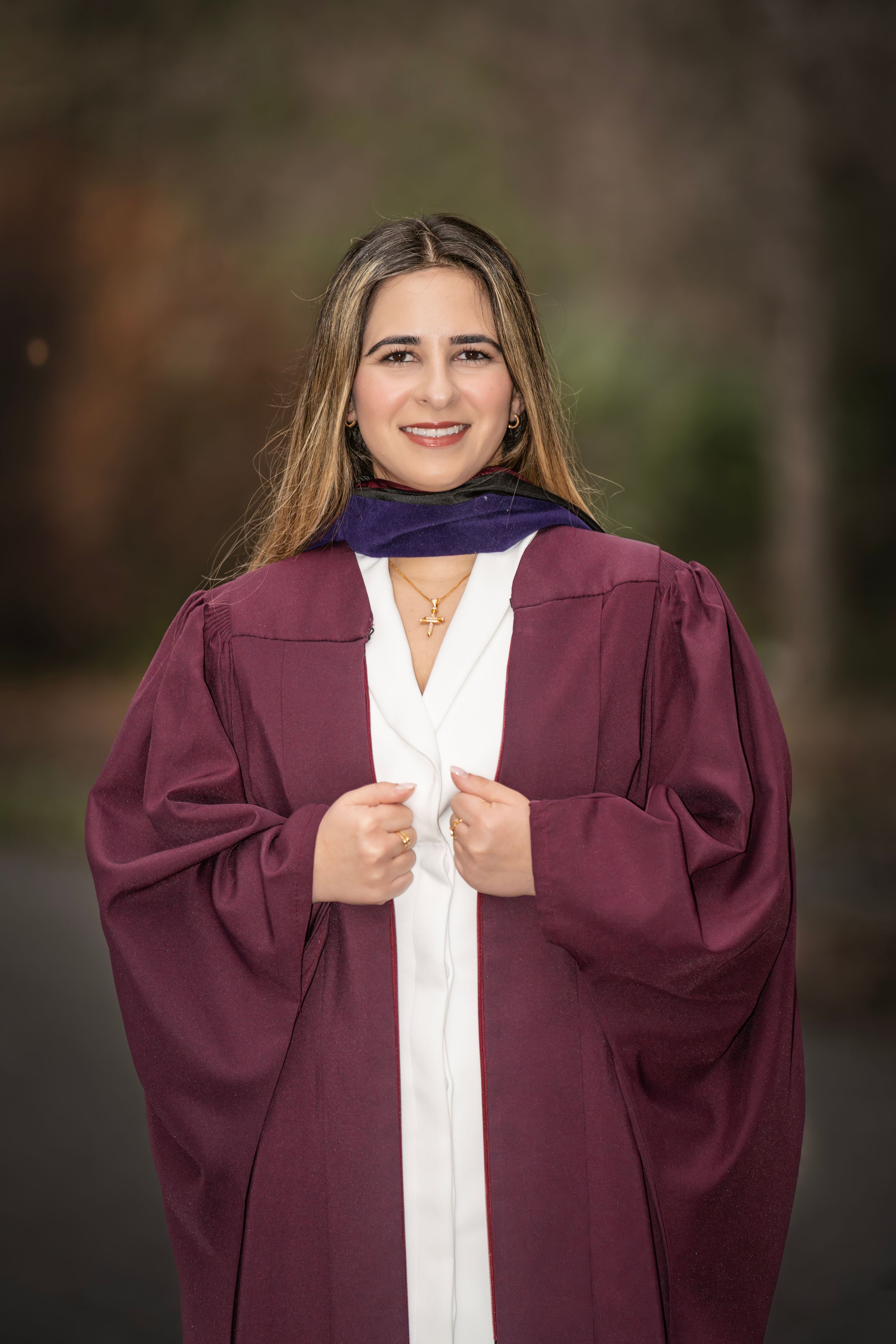 Smiling young woman in maroon graduation gown and navy hood poses outdoors for graduation portrait photo.