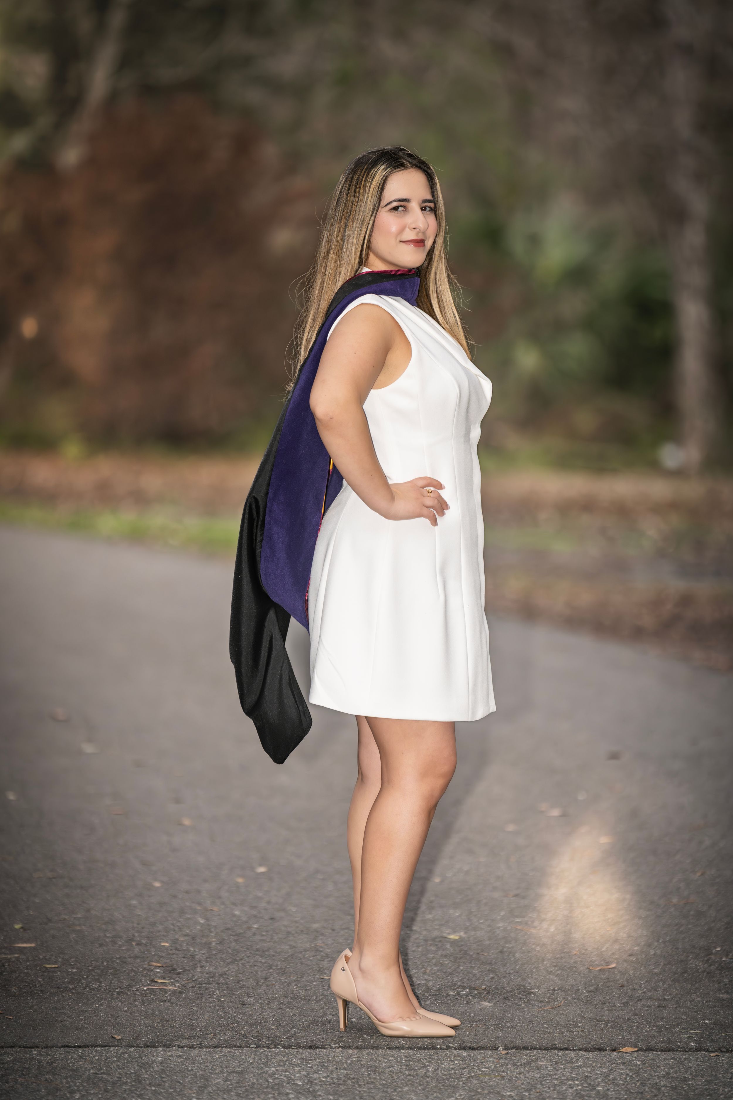 Young woman in white dress and heels holds graduation regalia while posing outdoors on a paved path.