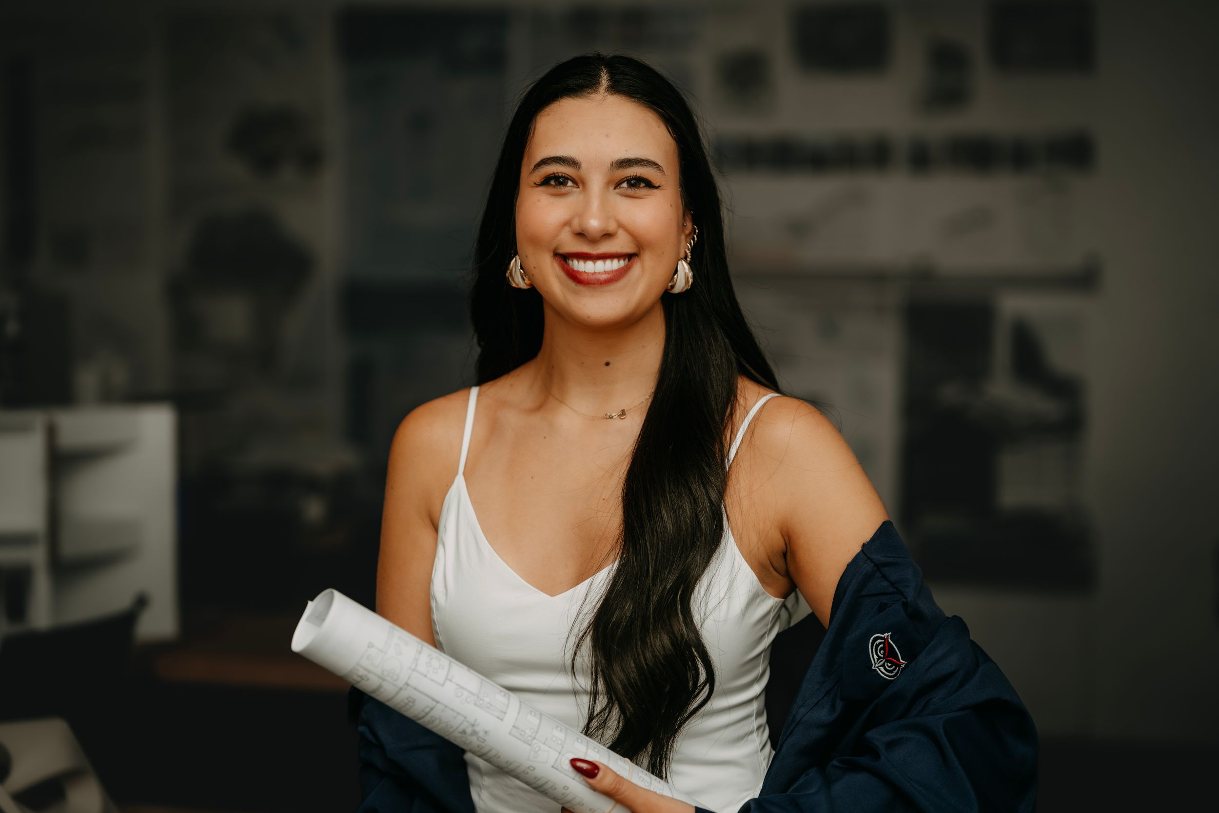 Smiling young woman holding rolled blueprints in a white top and blue jacket, standing in a modern office.