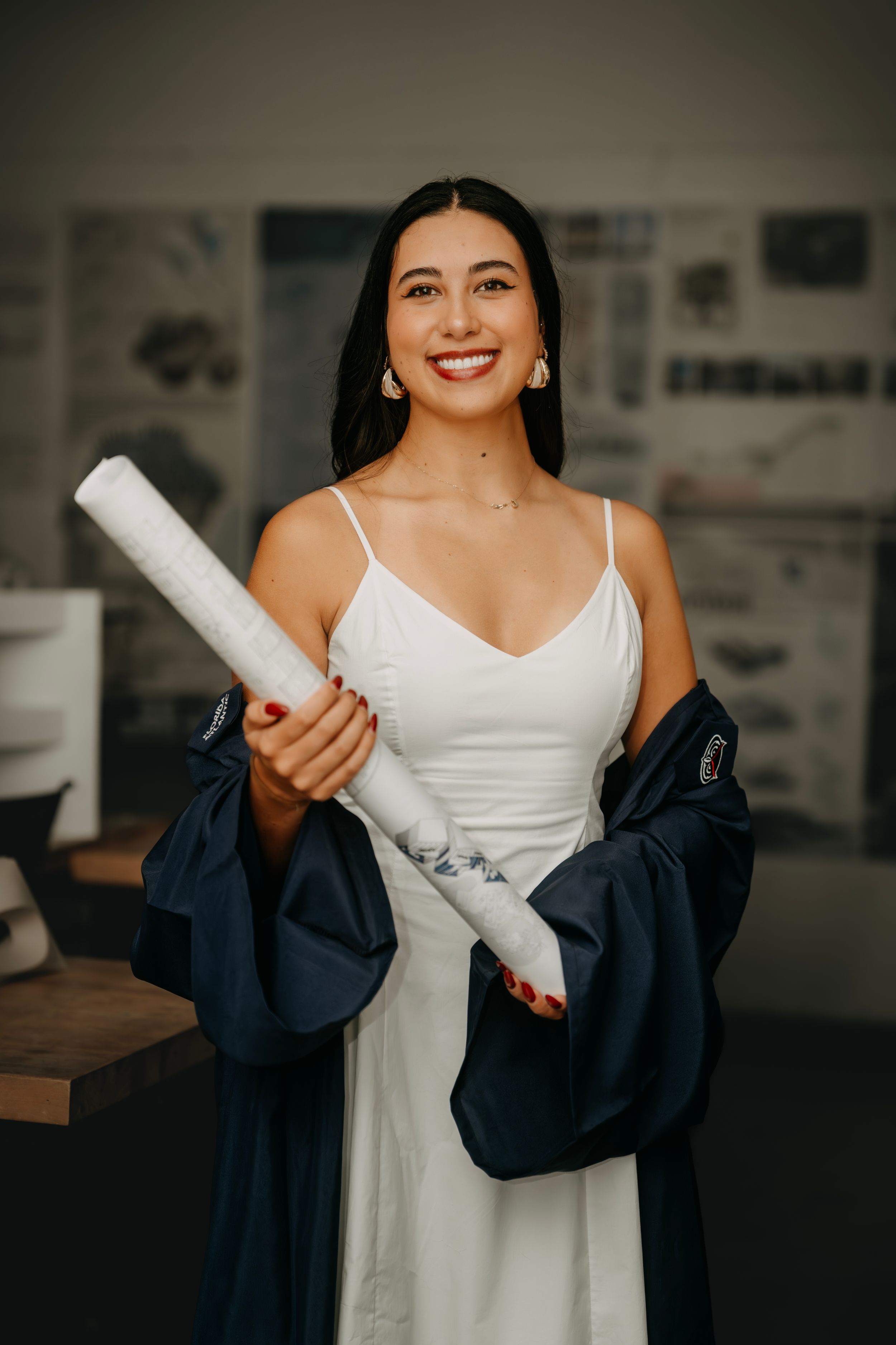 Smiling young woman in white dress and navy graduation gown holding a rolled diploma in a modern studio.