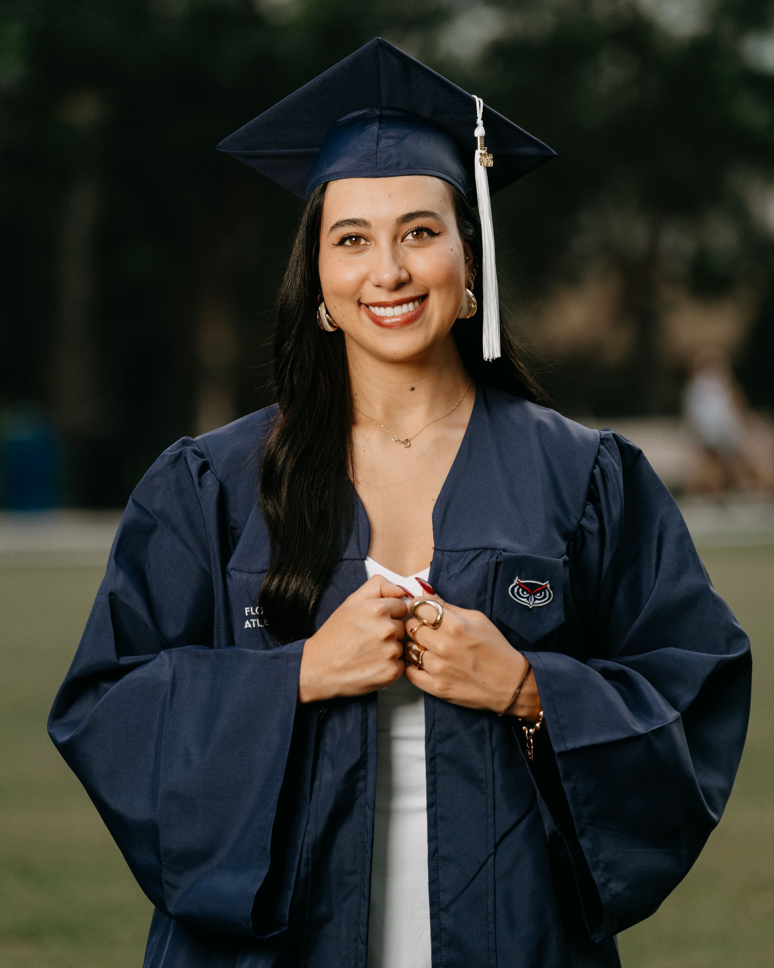 Smiling young woman in navy blue graduation cap and gown holding diploma outdoors on a green campus.
