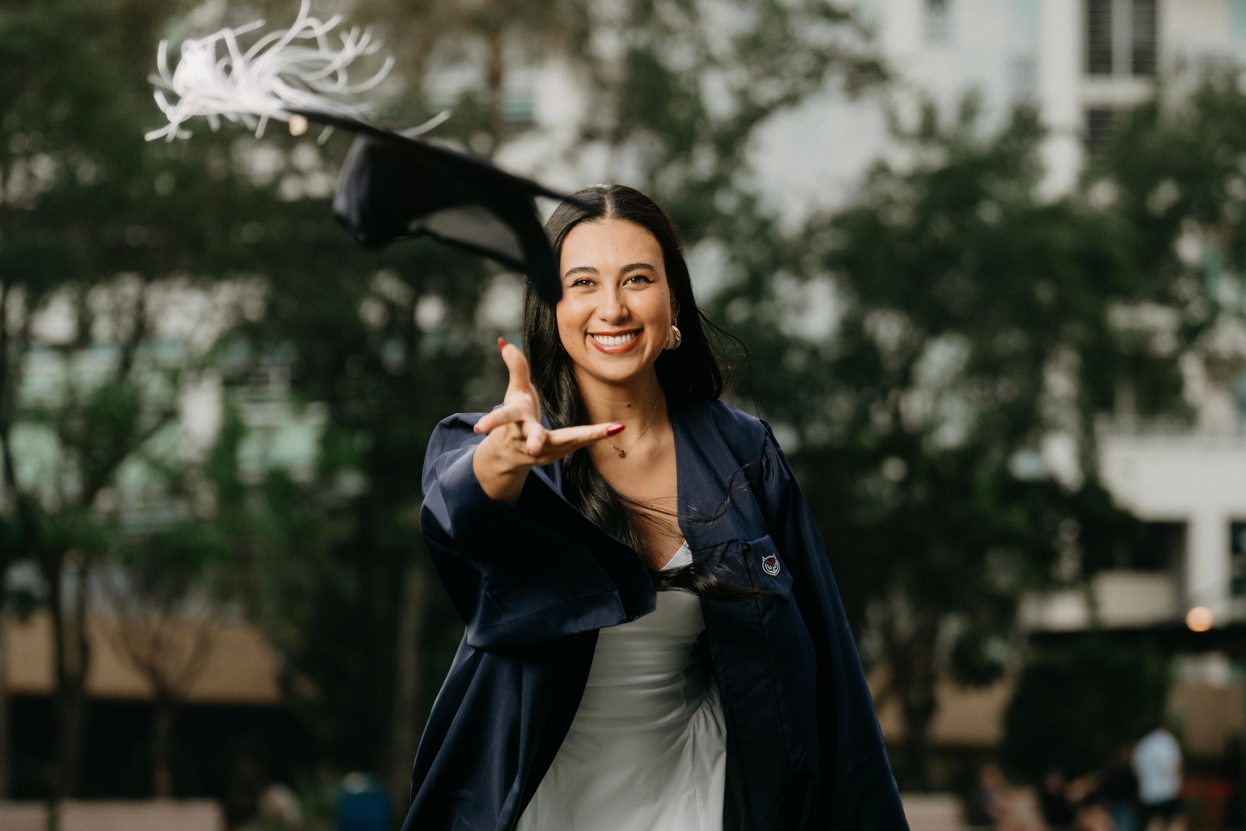 Smiling graduate in navy gown tossing cap in the air outdoors, celebrating academic achievement.