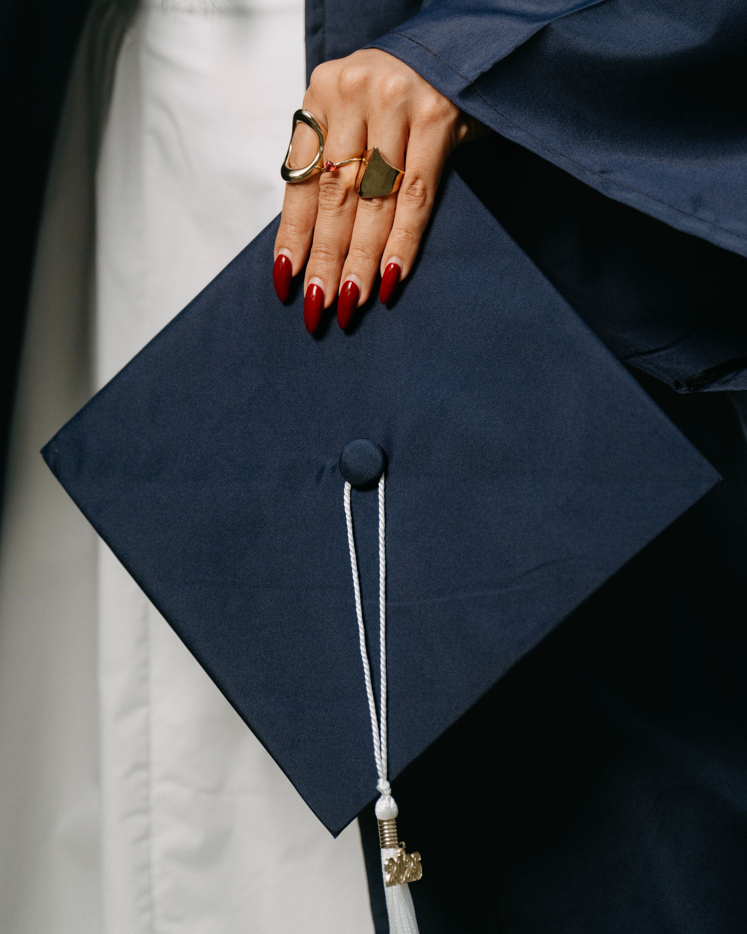 A graduate holds a navy blue mortarboard cap with white tassel, wearing gold rings and red nails.