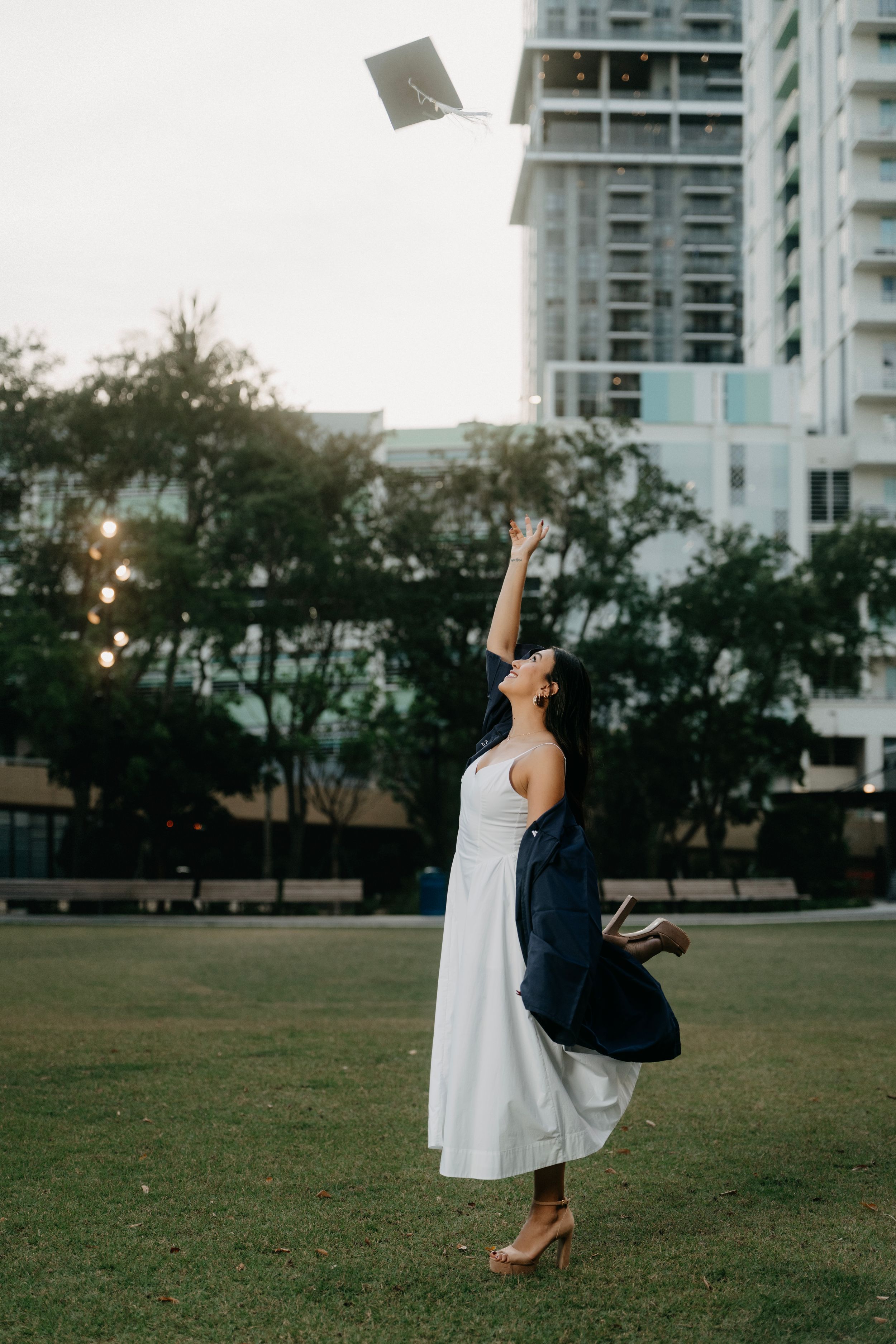 Graduate in white dress tosses cap in the air outdoors, celebrating her graduation achievement.