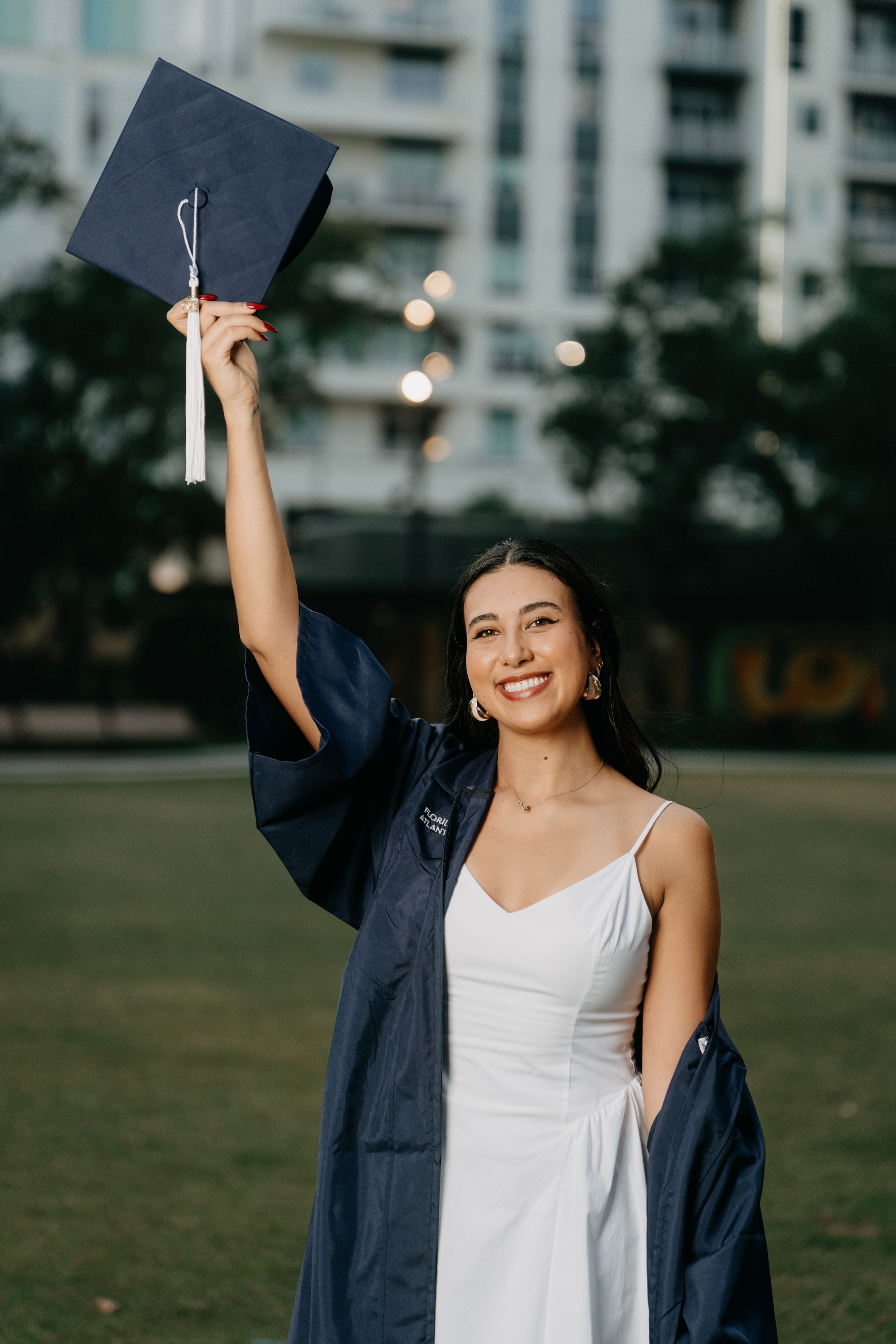 Smiling young woman in white dress and navy graduation gown holding cap up outdoors on campus green.