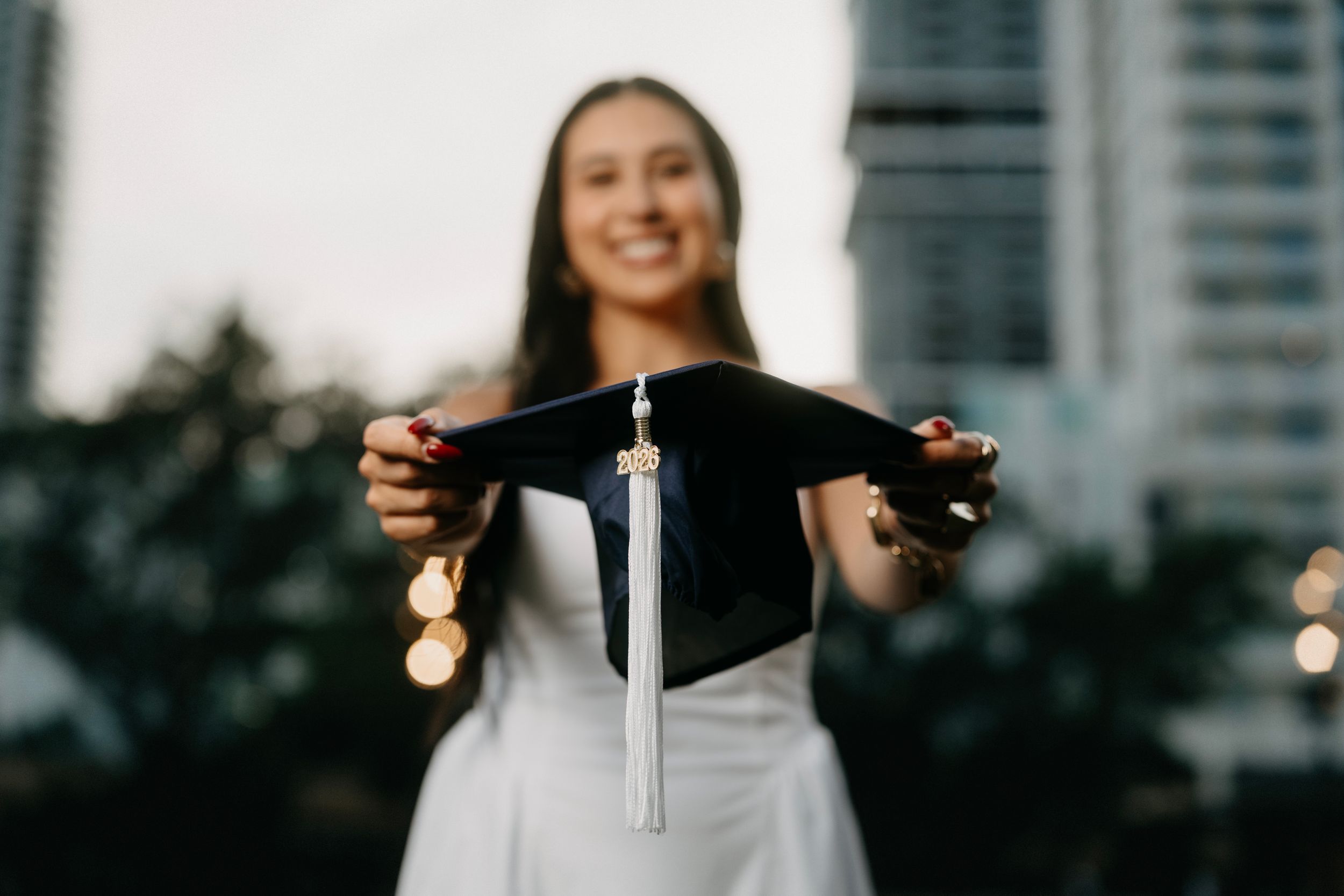 Smiling graduate holding a black mortarboard cap with white tassel outdoors at dusk in a white dress.