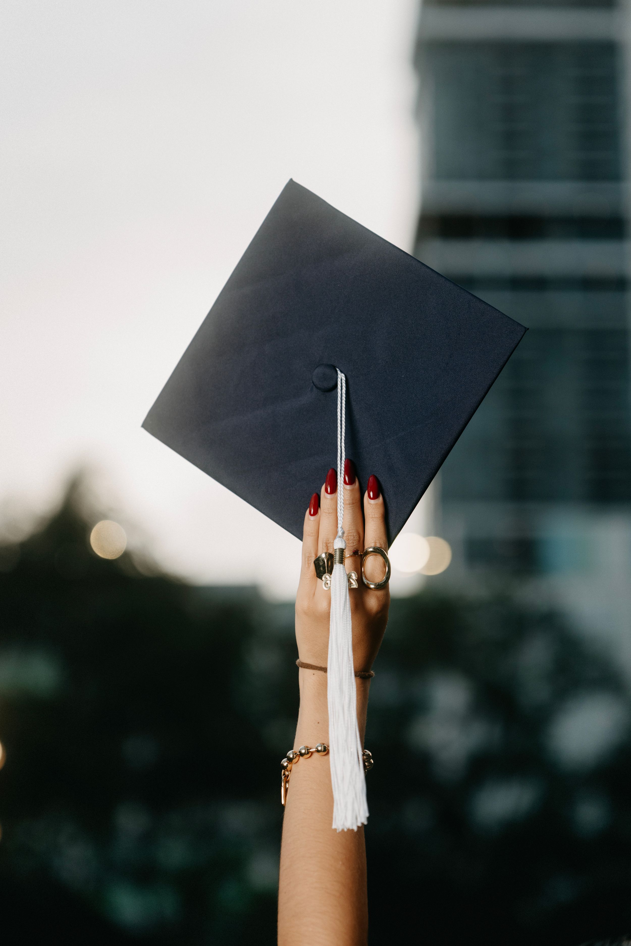 A hand with red nails and gold rings raises a black graduation cap with white tassel against a blurred urban background.