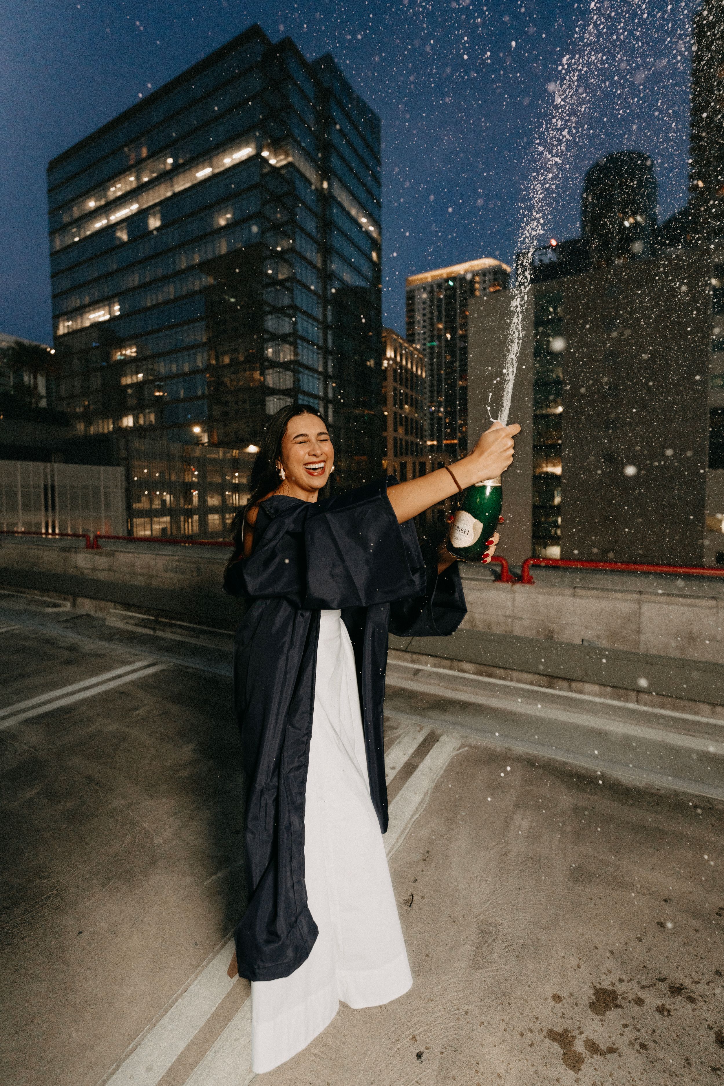 Graduate in cap and gown joyfully sprays champagne on rooftop at night with city buildings lit up behind her.