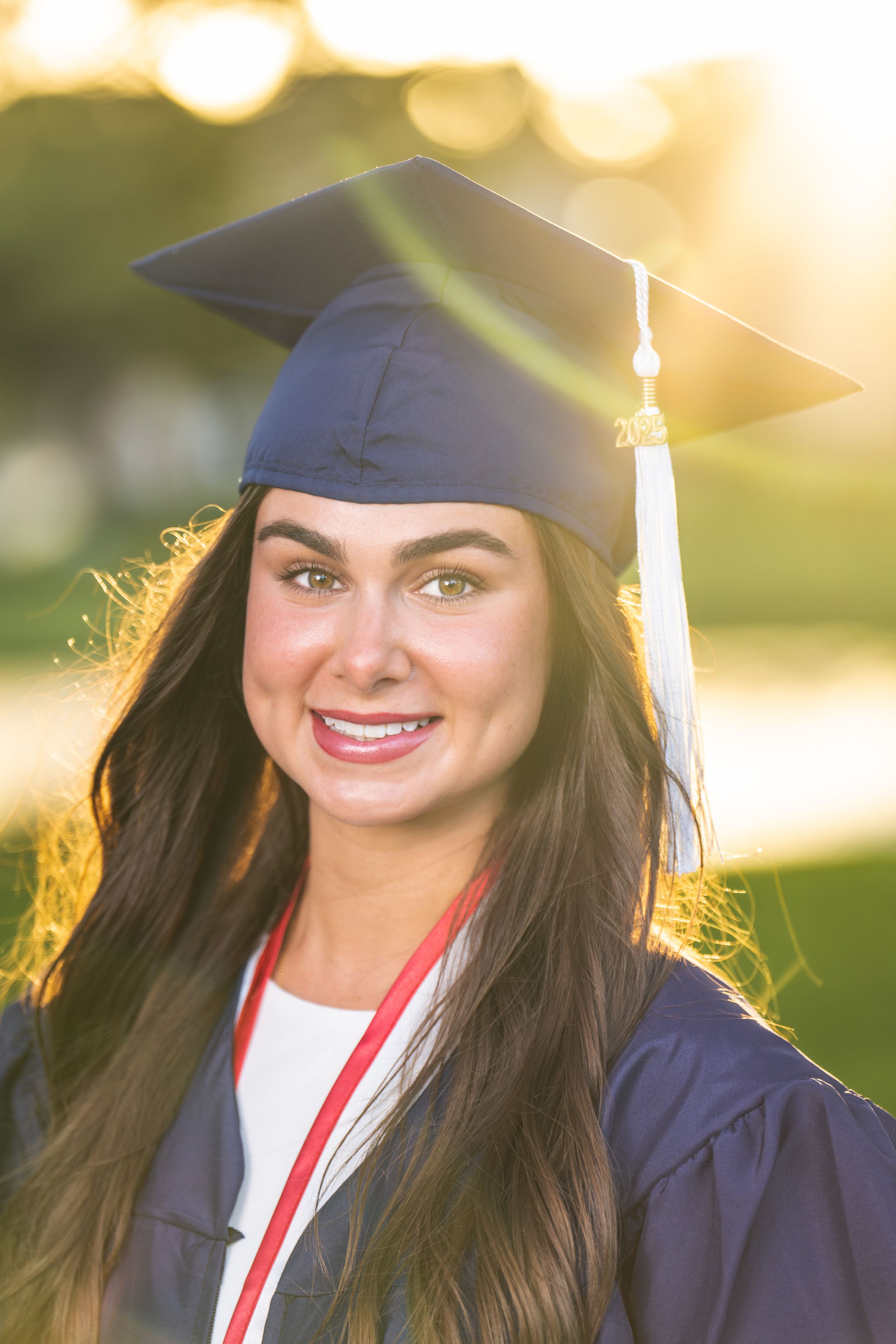 Smiling young woman in navy blue graduation cap and gown posing outdoors in golden sunlight.