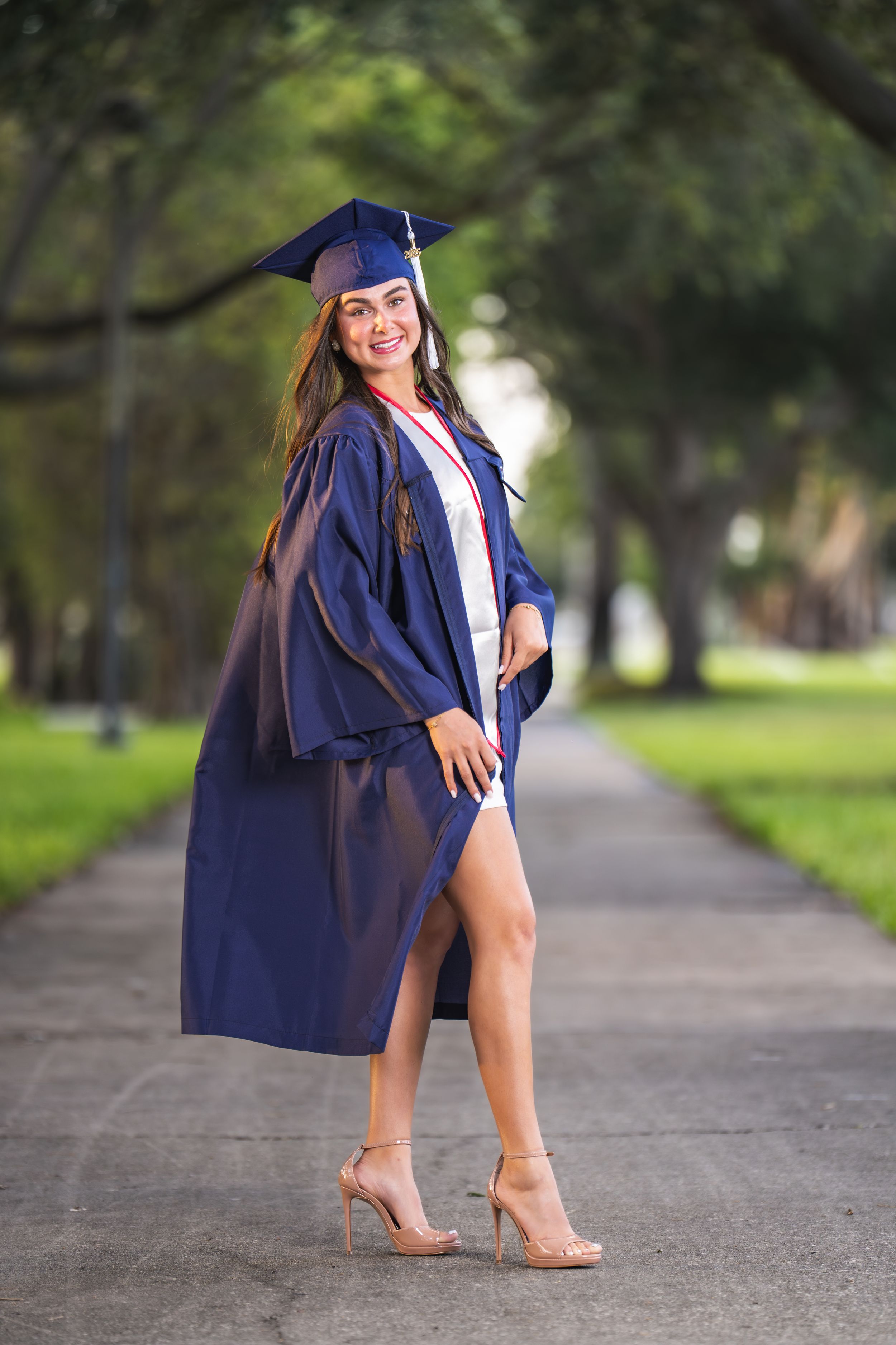 Smiling female graduate in navy blue cap and gown posing on a tree-lined campus walkway wearing heels.