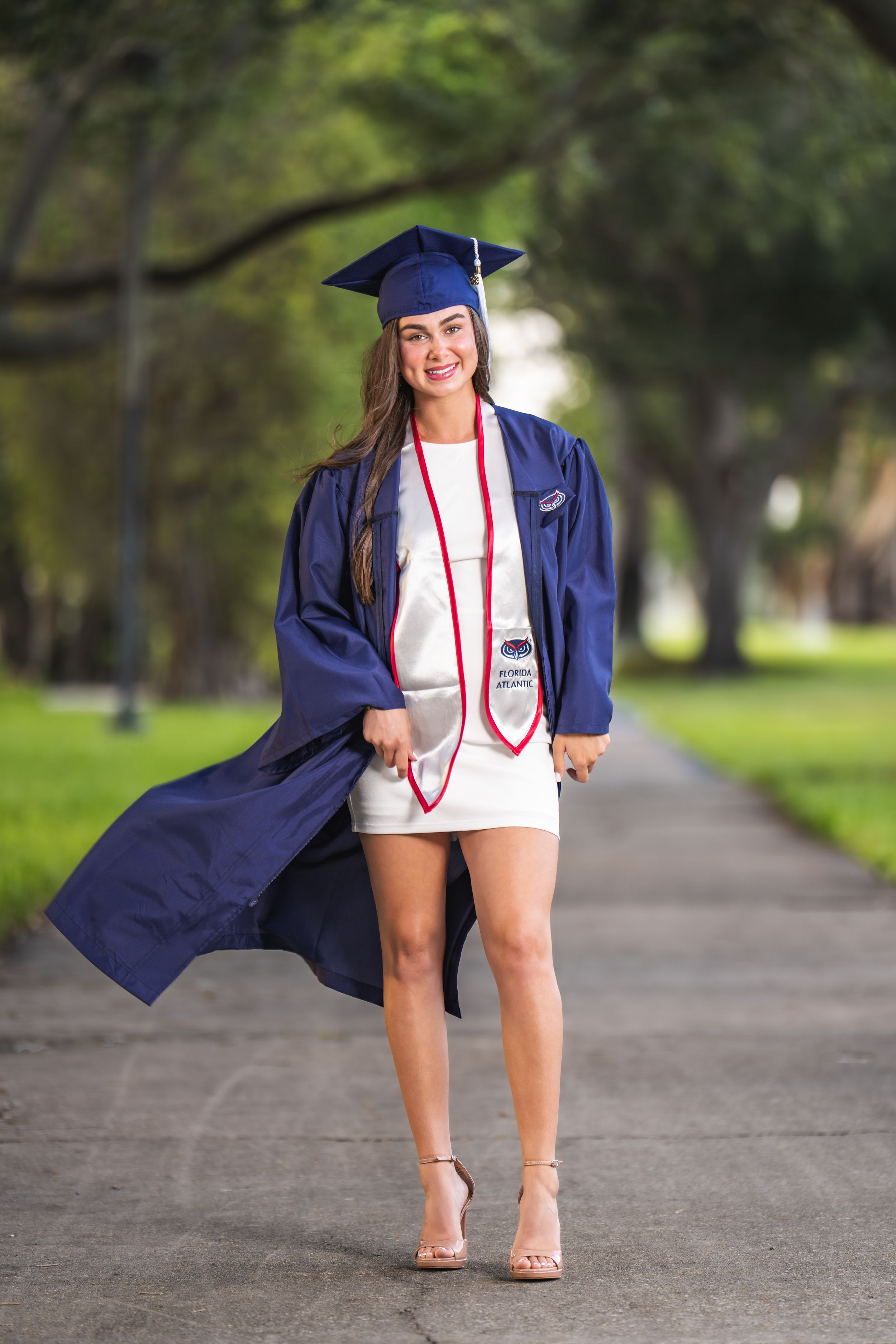 Smiling young woman in navy blue graduation cap and gown posing on a tree-lined pathway in heels.