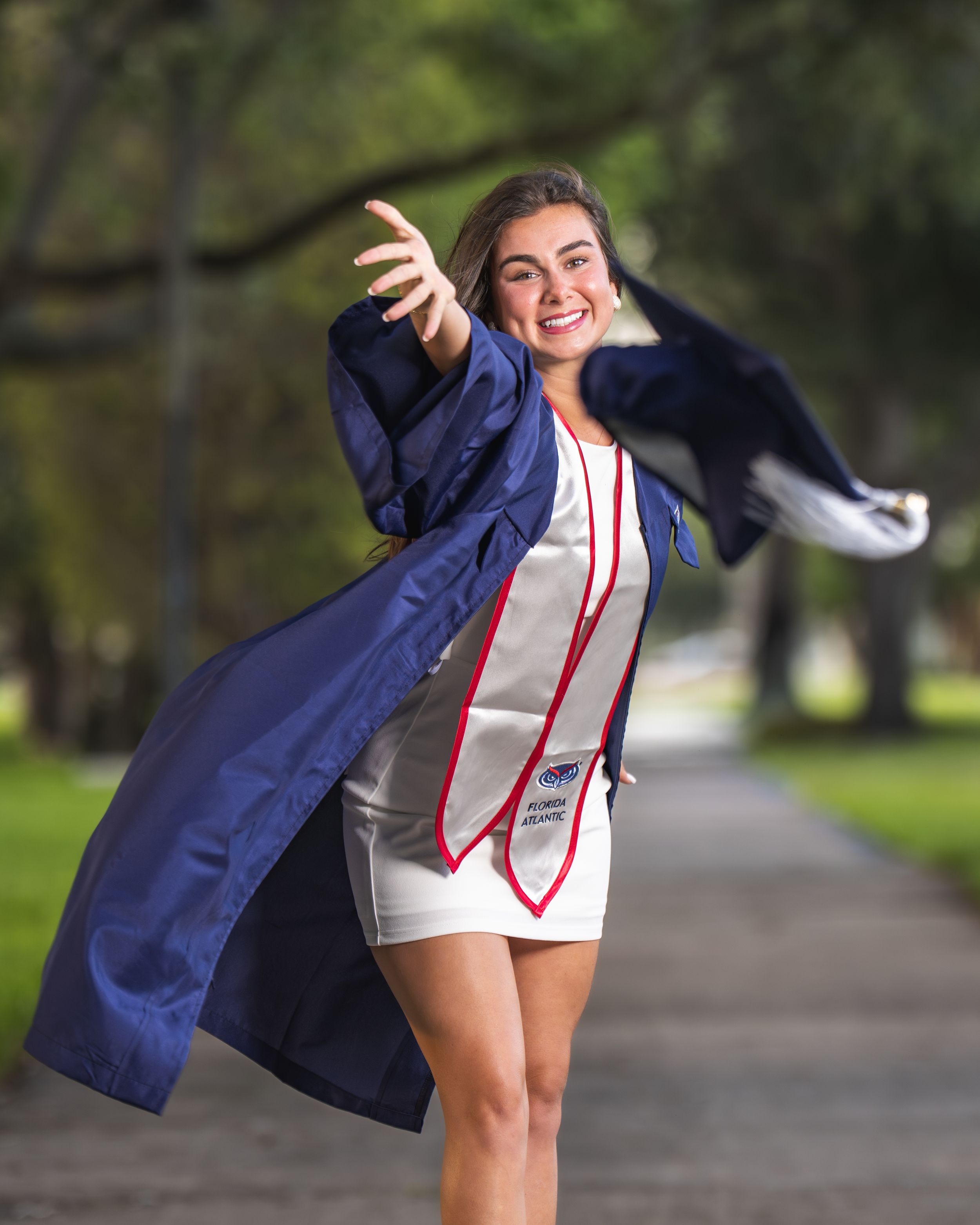Smiling graduate in navy blue cap and gown tossing her robe outdoors on a tree-lined path.