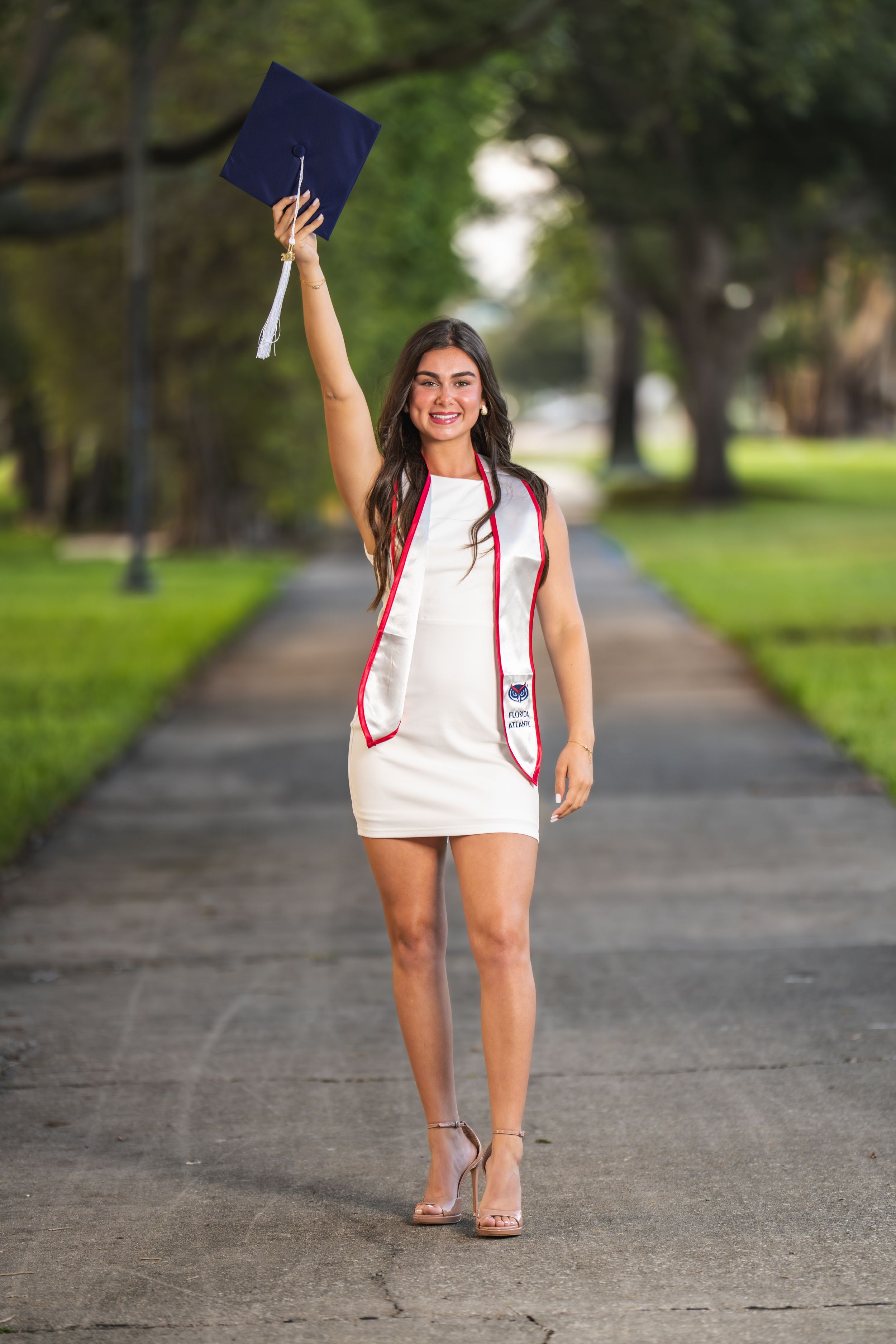 Young woman in white dress holding graduation cap aloft on a tree-lined path, celebrating her academic achievement.